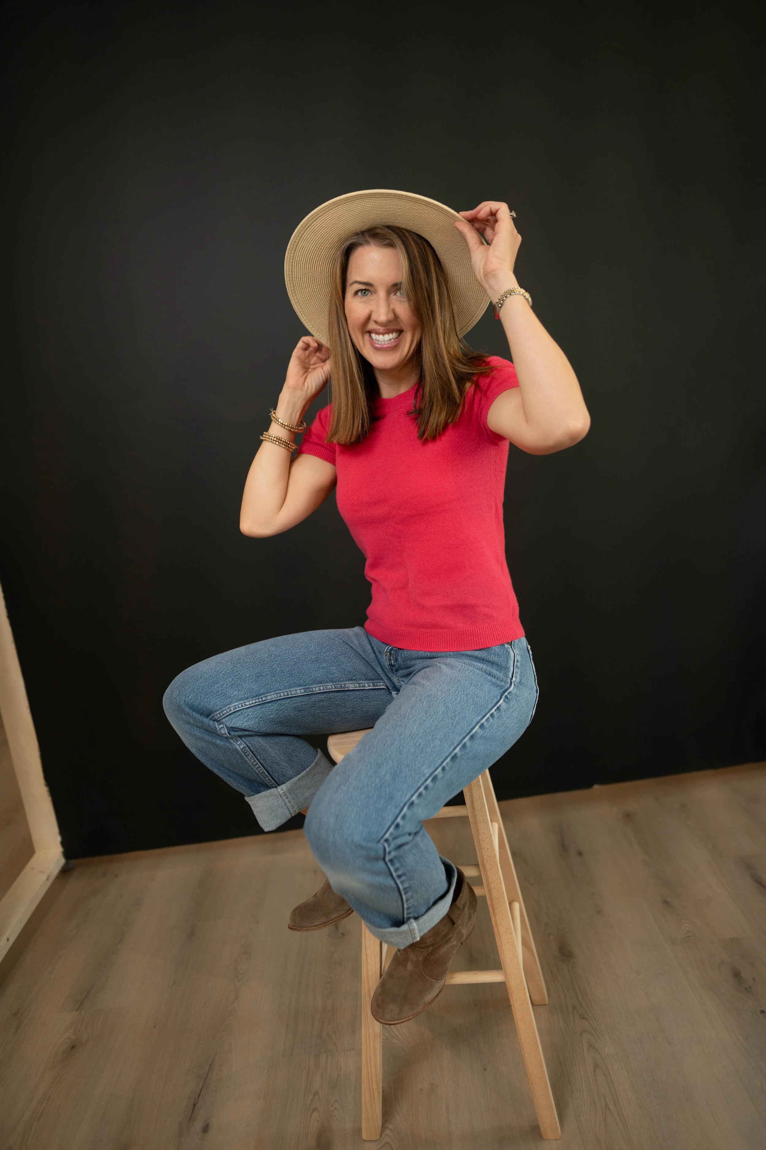 A woman sitting on a tall wooden stool, wearing a red shirt, blue jeans, and brown boots, holding a wide-brimmed straw hat with one hand, smiling, in front of a dark backdrop.
