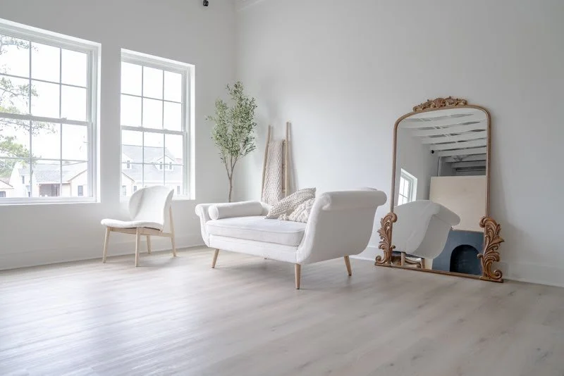 Minimalist living room with large windows, white walls, light wood flooring, a white armchair, a white sofa with cushions, a tall green plant, a decorative floor lamp, and an ornate mirror leaning against the wall.