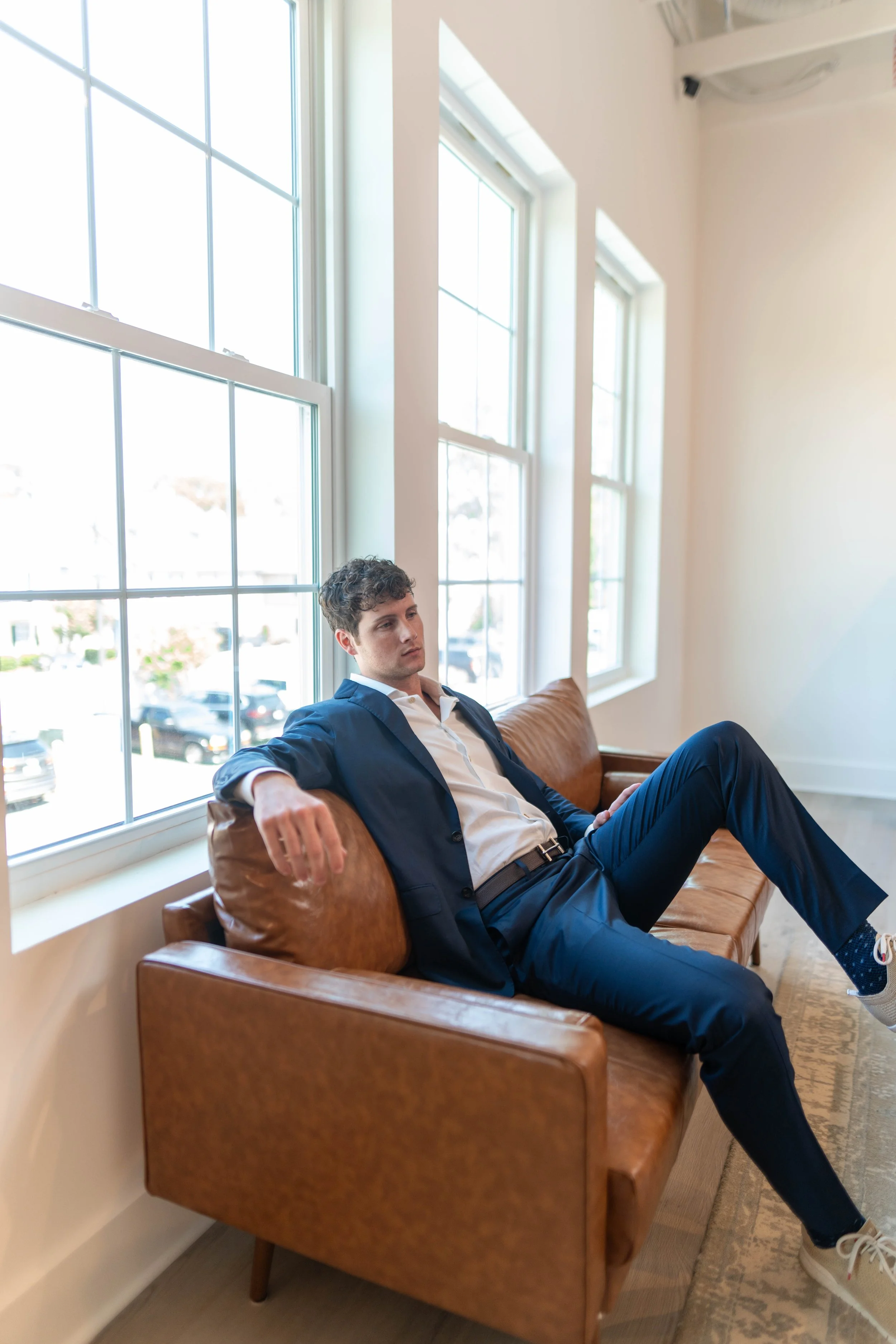 A young man in a navy suit and white shirt is sitting on a brown leather couch near large windows, with sunlight coming in.