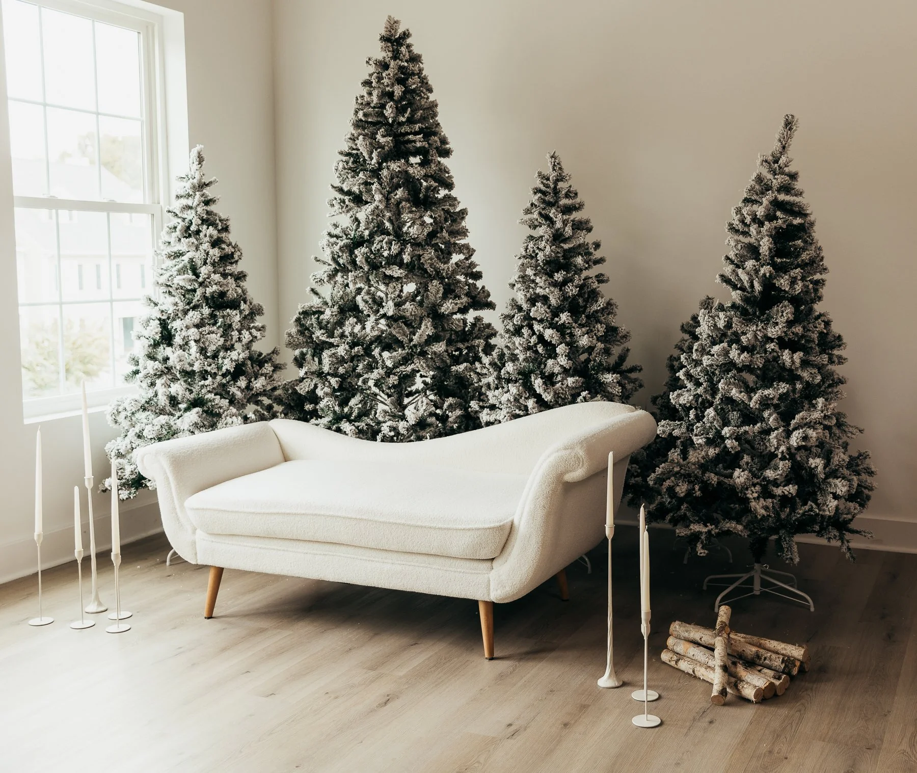 Decorative winter scene indoors with four snow-dusted Christmas trees, a white vintage sofa, white candlesticks, and a small pile of logs near a window with white walls and light wooden flooring.