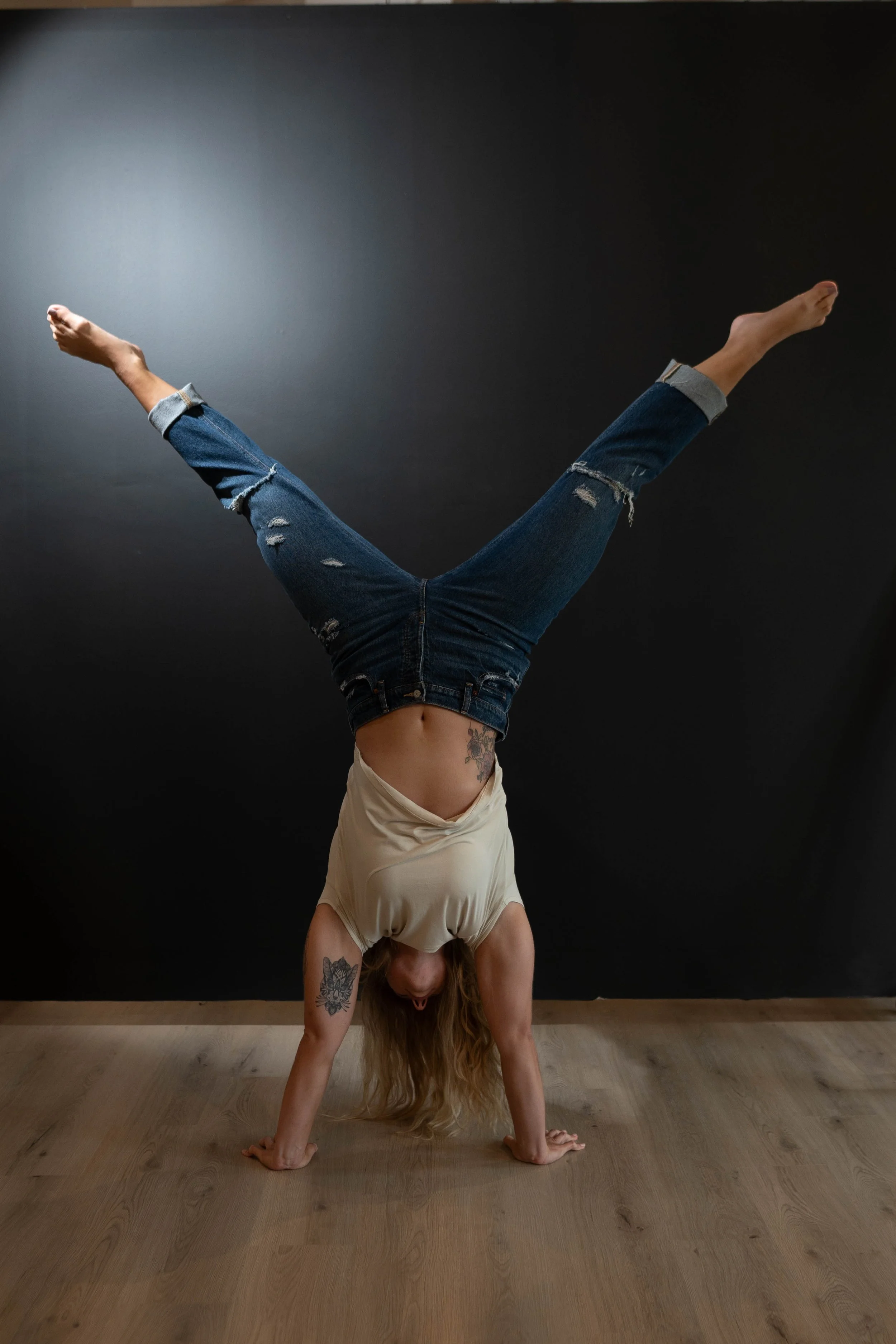 Person performing a handstand with legs spread in a V shape in a room with gray walls and wooden floor.