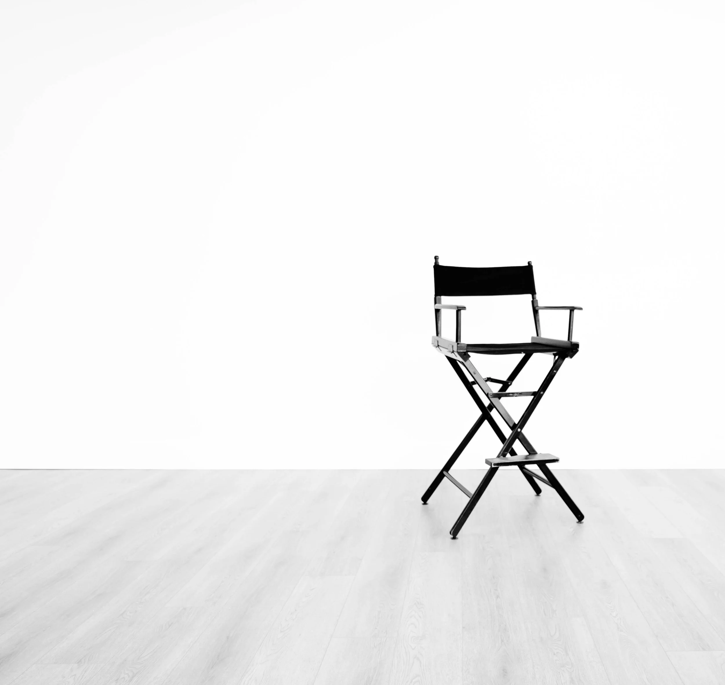 A black director's chair with wooden armrests and a black fabric seat and backrest positioned on a white wooden floor against a plain white wall.