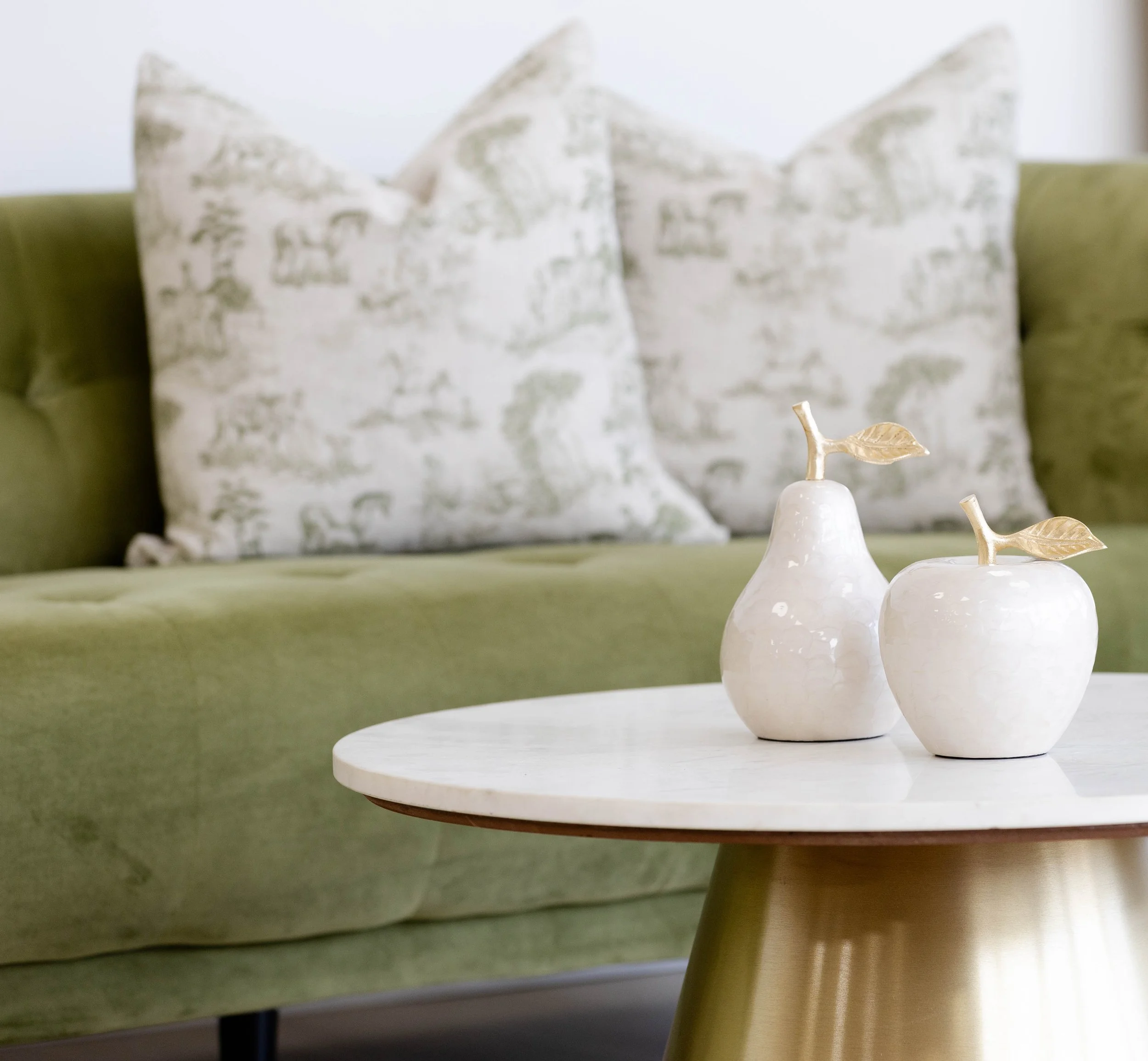 A close-up of two small white ceramic vases with gold-colored leaves on a white circular tray, placed on a green surface, with a background of decorative pillows.