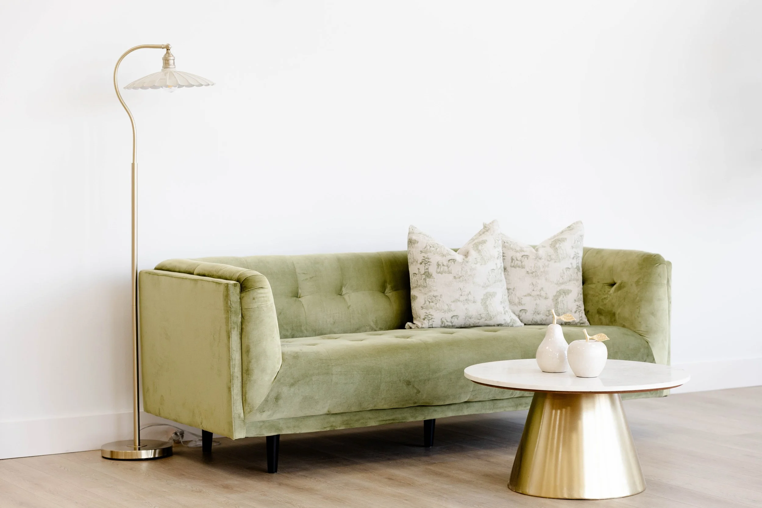 A light green velvet sofa with tufted backrest, two patterned pillows, a round white marble-top coffee table with a gold base, two white decorative vases, and a gold floor lamp with a white scalloped lampshade, against a plain white wall.
