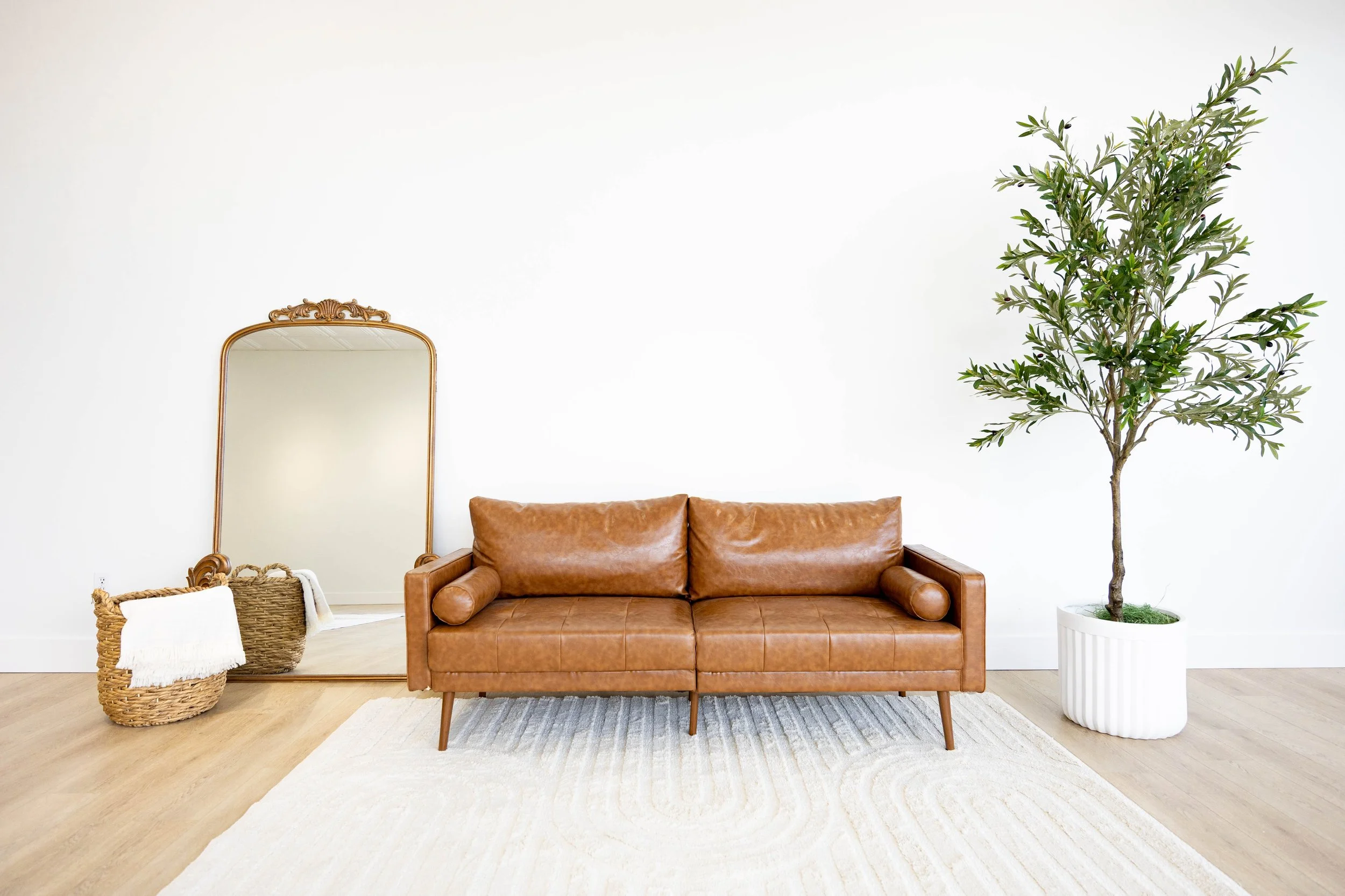 Living room with a brown leather sofa, a white rug, a large potted tree, a woven basket with a throw blanket, a vintage mirror, and a neutral wall.