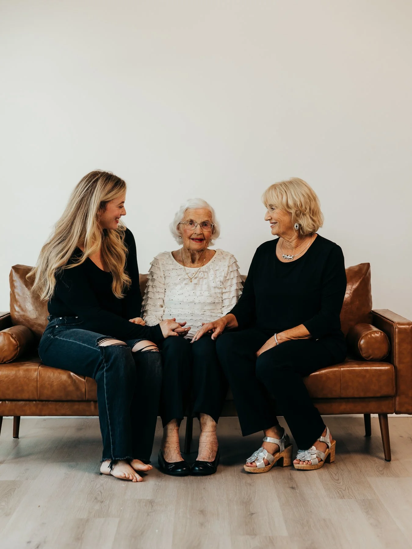 Three women sitting on a brown leather couch, engaged in conversation and smiling. The women appear to be of different generations, with the older woman in the middle and the younger women on each side. The setting is a simple, modern room with plain