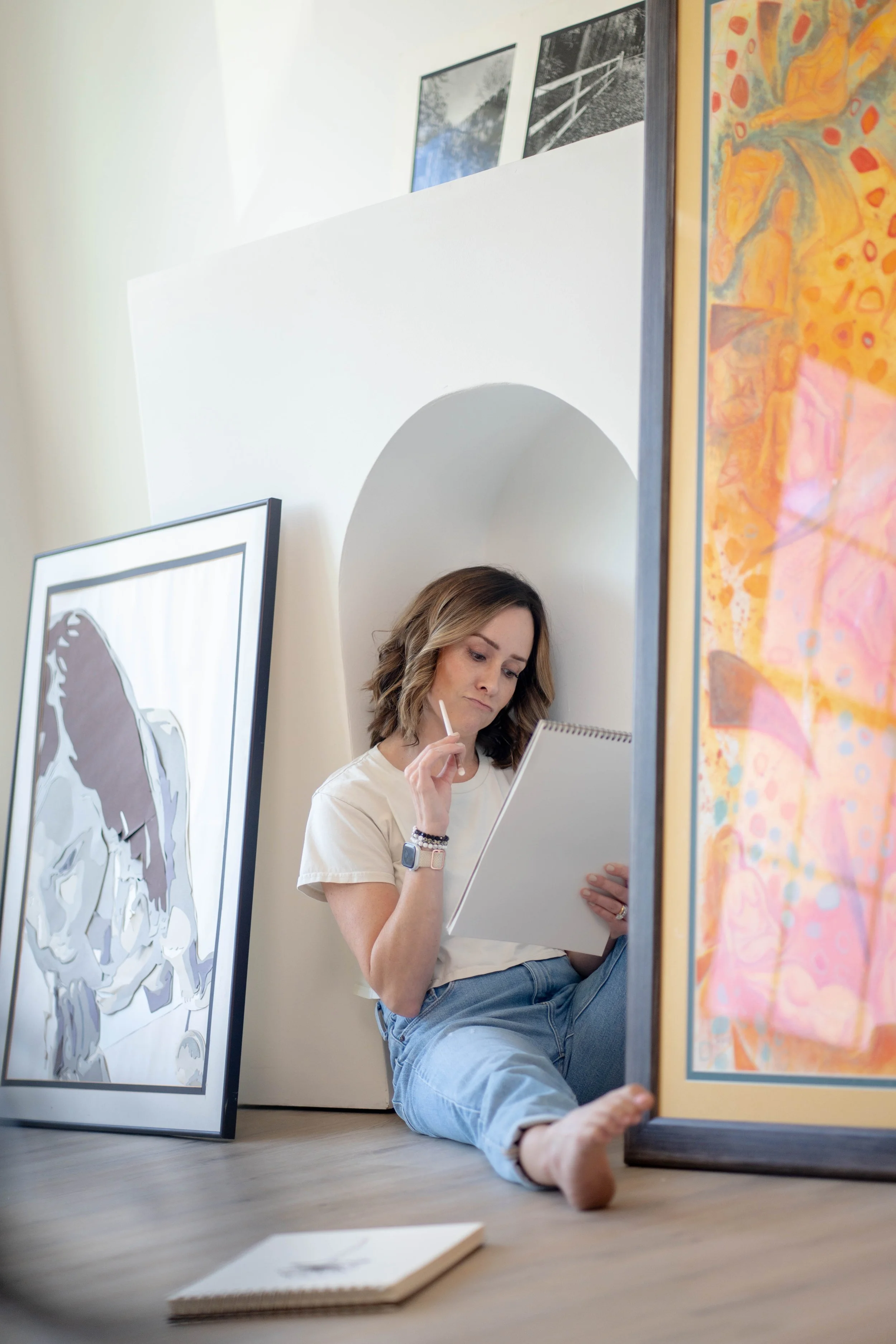 A woman in a white shirt and jeans is sitting on the floor, leaning against a large framed painting, holding a notepad and drawing with a pencil. There are other artworks on the wall and floor behind her, and she is surrounded by art supplies and fra
