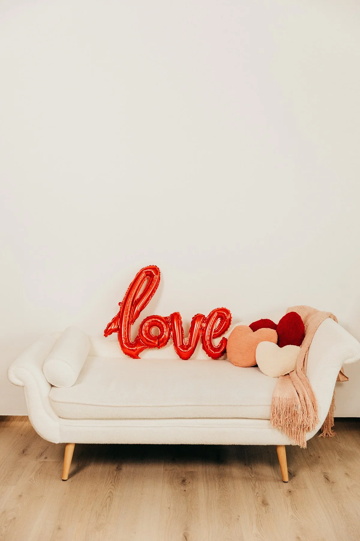 A white sofa with a rolled armrest and wooden legs, decorated with a pink fringed blanket and fuzzy heart-shaped pillows, with a red balloon spelling 'love' and plush heart cushions on top, against a plain white wall.