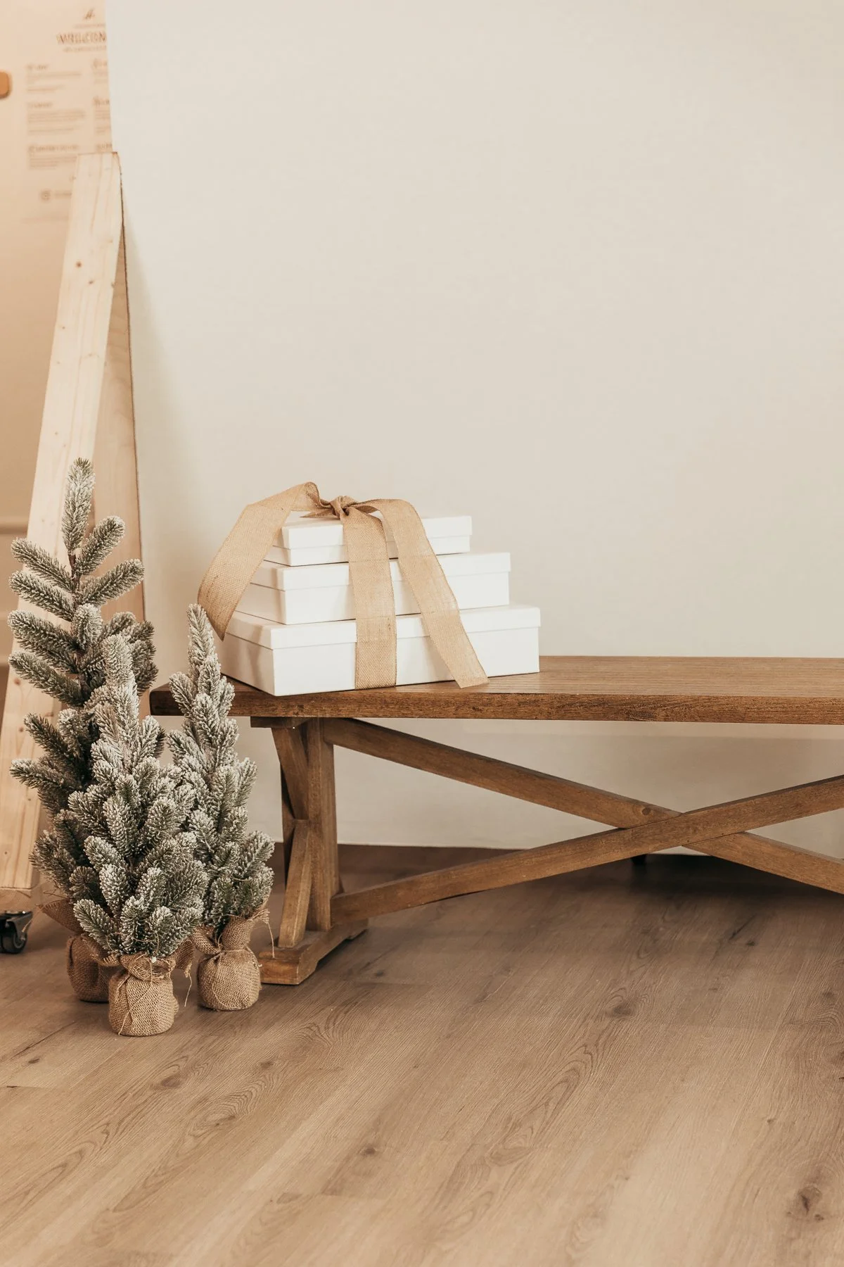 Small potted fir tree with burlap sack at base, placed next to a wooden table. On the table, there are four white gift boxes stacked and tied with a beige ribbon. A wooden ladder and a beige wall with a faintly visible poster are in the background.