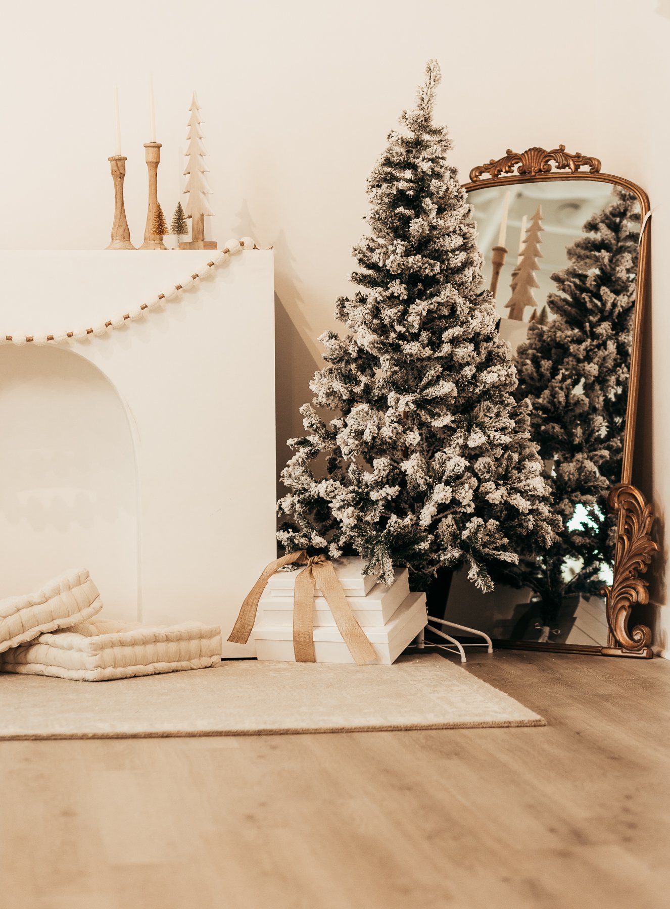 A decorated Christmas scene with a Christmas tree covered in faux snow, a large mirror reflecting the scene, wrapped gifts underneath the tree, and a cozy blanket on a beige rug.
