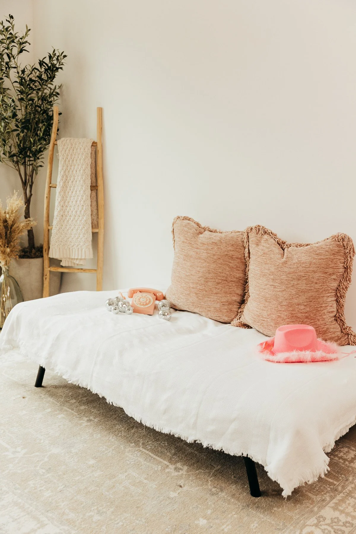 A white sofa with two pink-fringed pillows on a beige carpeted floor. On the sofa, there is a vintage pink rotary phone and a string of silver Christmas ornaments. To the left, a wooden ladder with a cream-colored knit blanket draped over it stands n