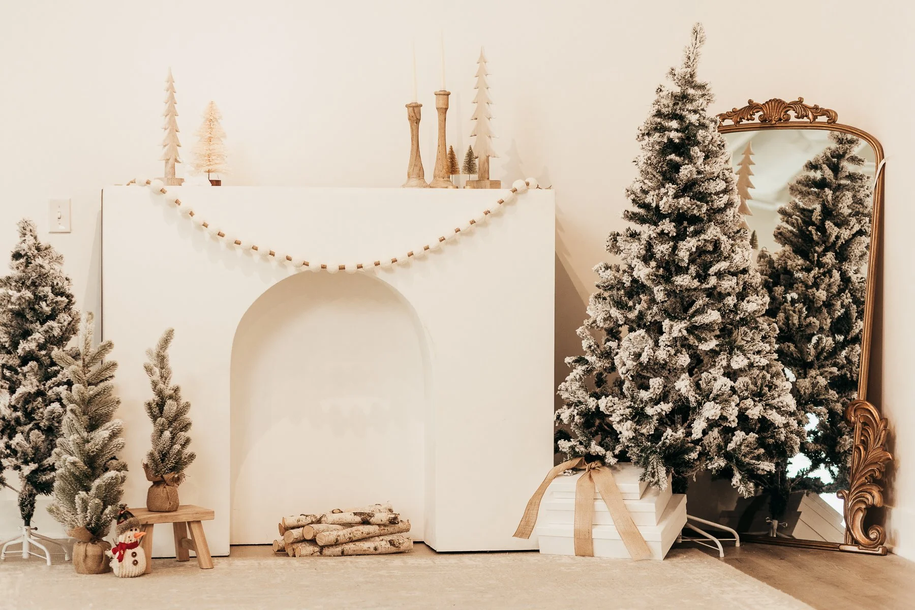 Decorated Christmas scene with snow-covered artificial trees, a white fireplace mantel adorned with small trees and candles, a small snowman, and a large vintage mirror reflecting part of the scene.