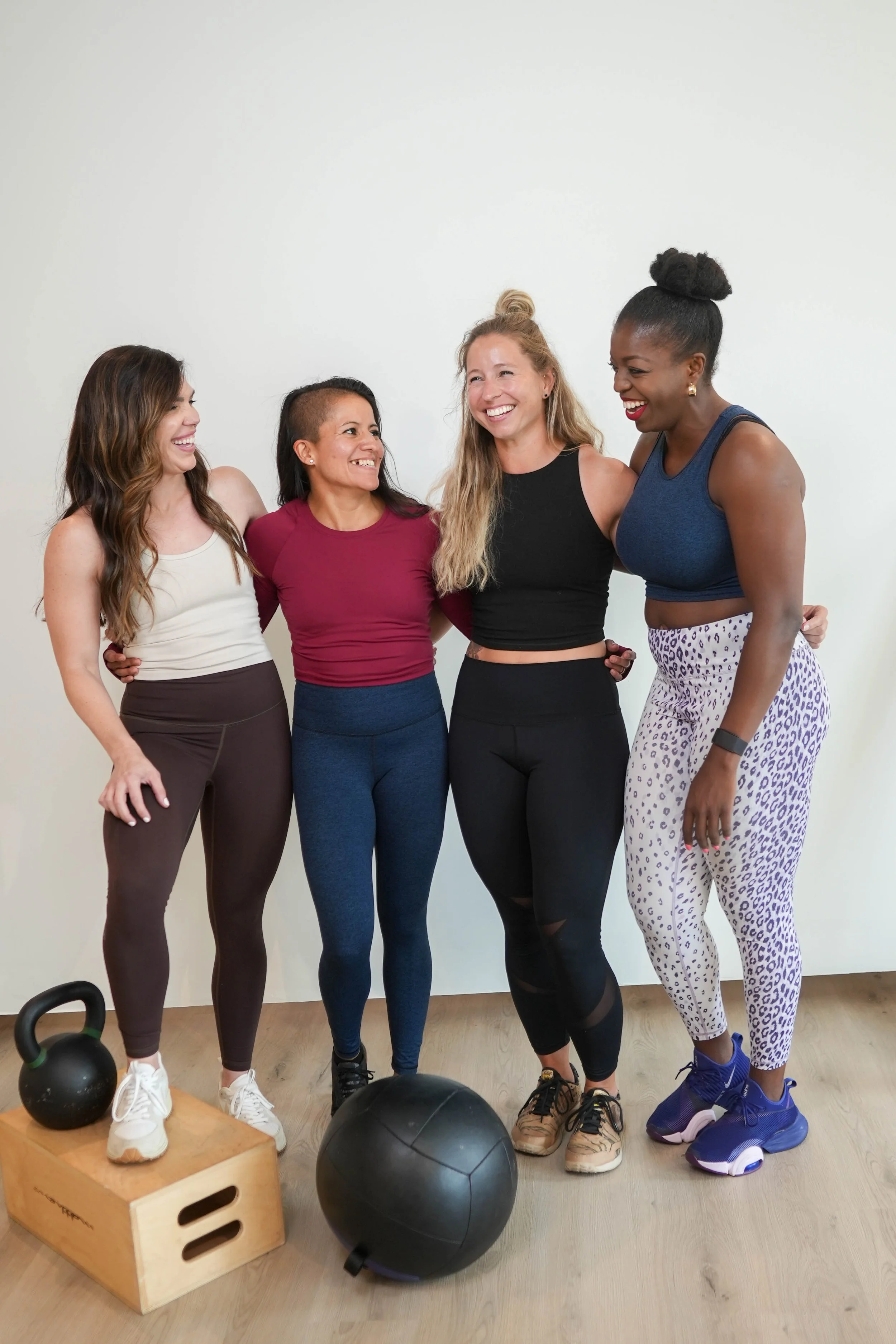 Group of four women in workout attire laughing and standing together in a gym with kettlebells and a medicine ball on the floor.