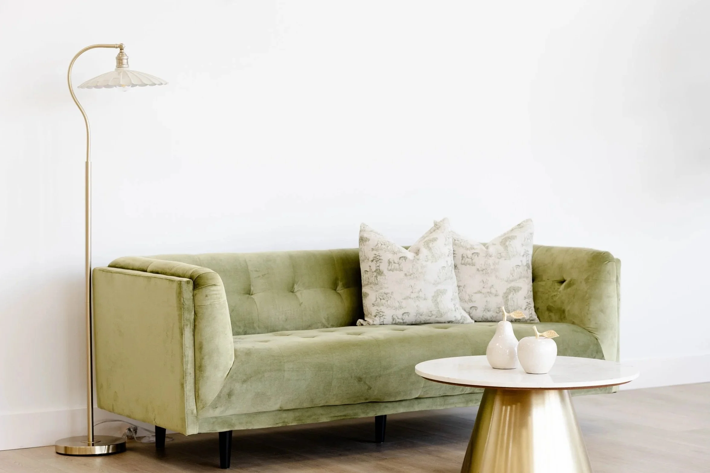 A green velvet sofa with three decorative pillows and a small white marble coffee table with vases. A tall brass floor lamp with a white shade stands beside it against an all-white wall.