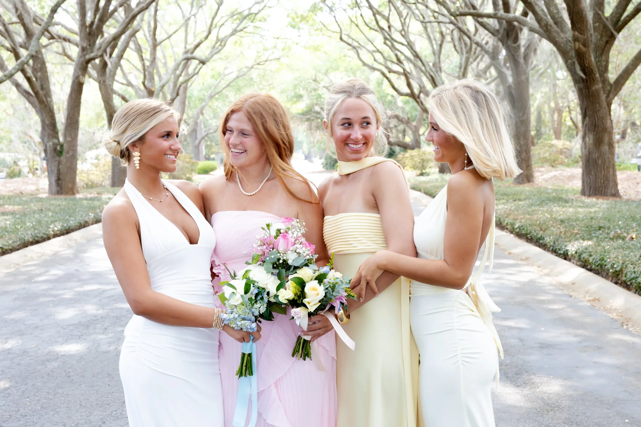 Four women dressed in pastel and white dresses, smiling and standing close together outdoors under tree-covered arches, with one woman holding a bouquet of flowers.