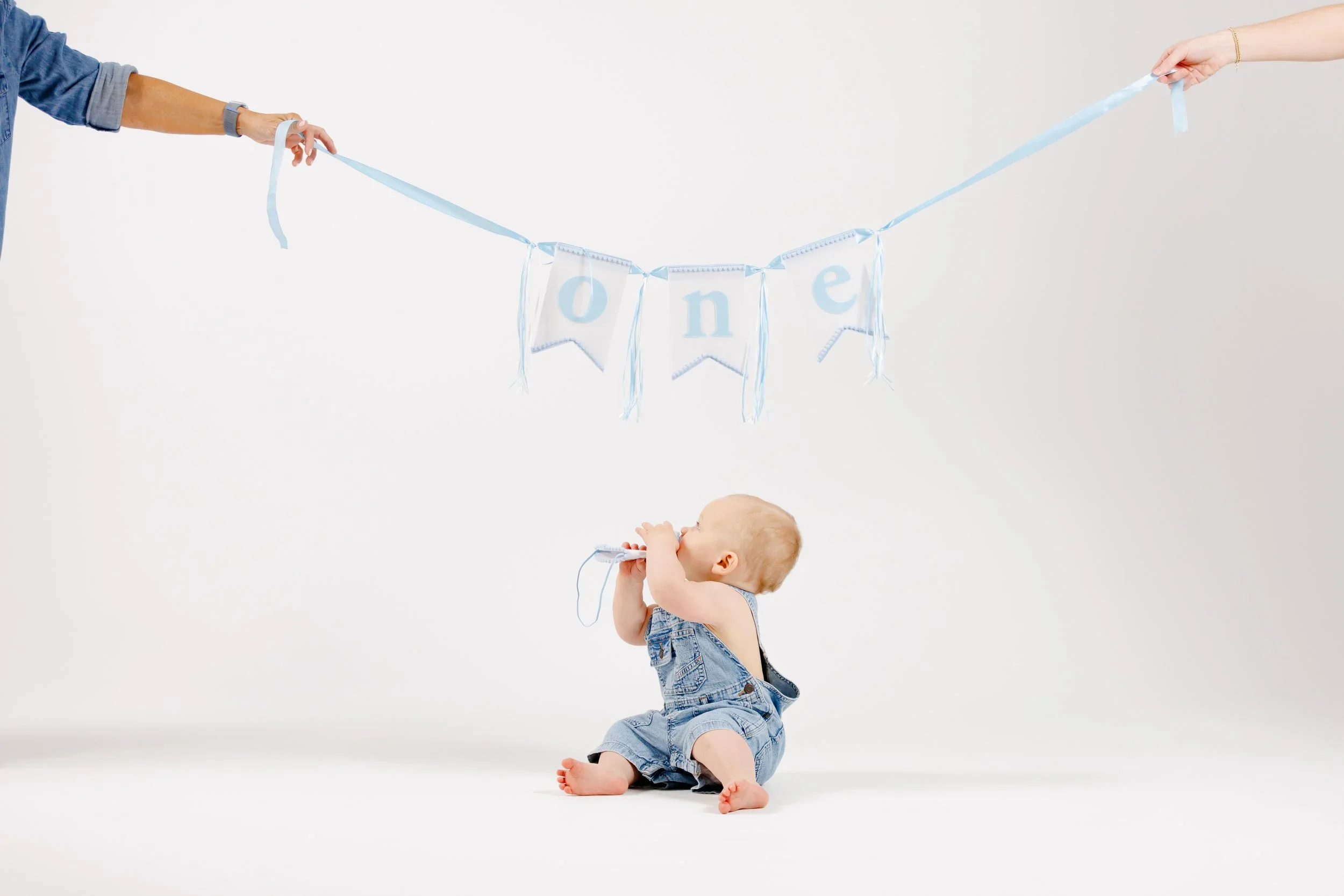 A young child sitting on the floor holding a blue ribbon with the word 'one' in light blue letters, with two hands holding each end of the ribbon.