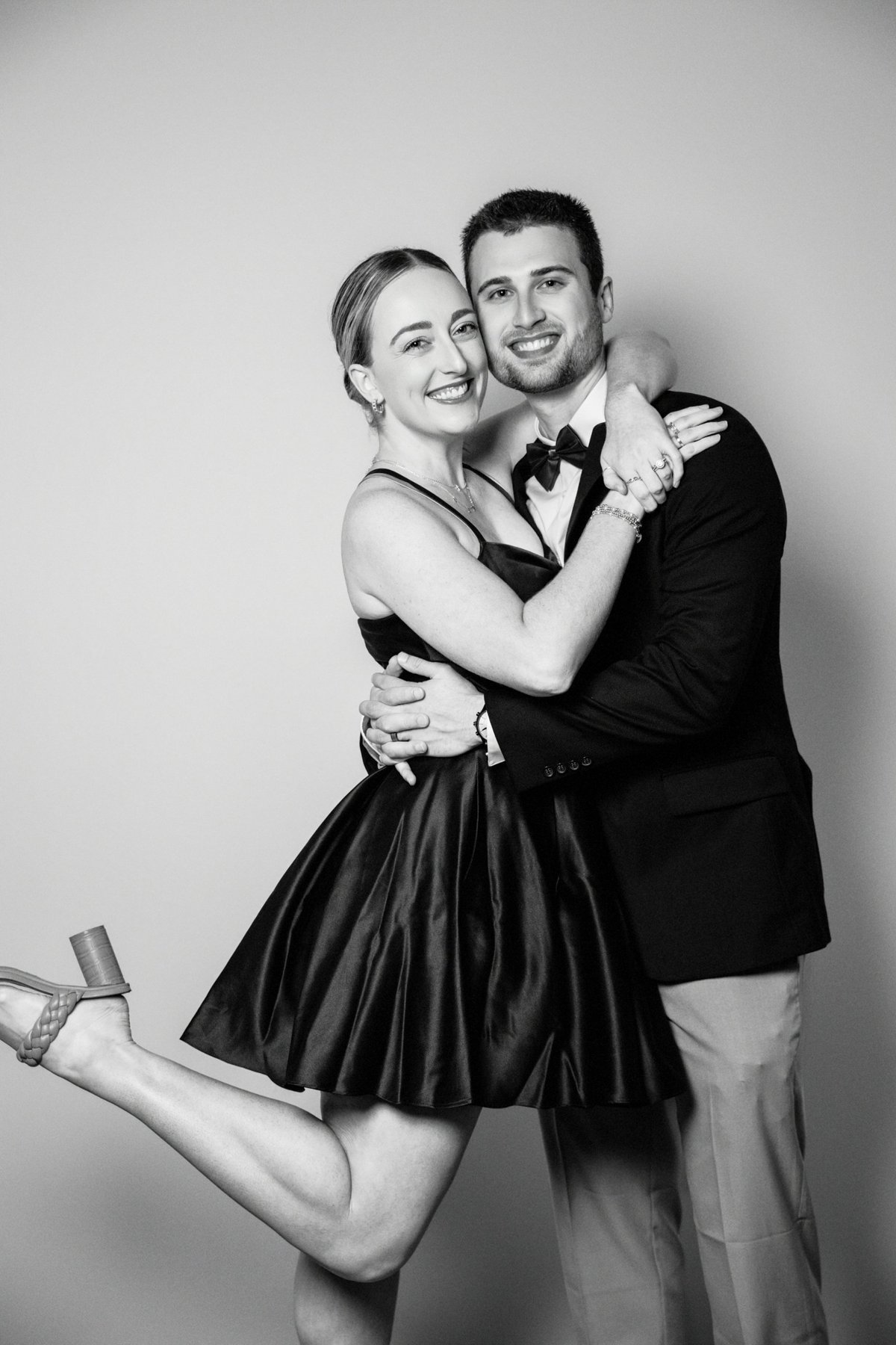 A black and white photo of a smiling couple dressed formally, with the woman in a sleeveless dress and high heels, and the man in a tuxedo, embracing each other against a plain wall.