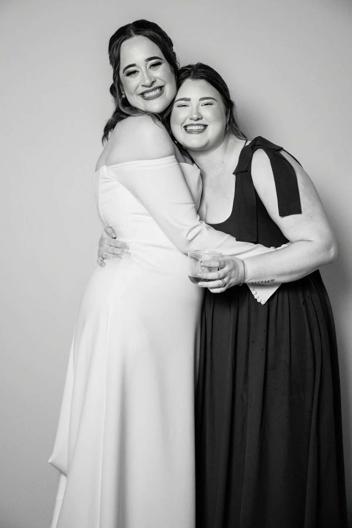 Two women smiling and hugging, one in a white dress and the other in a black dress, standing against a plain backdrop.