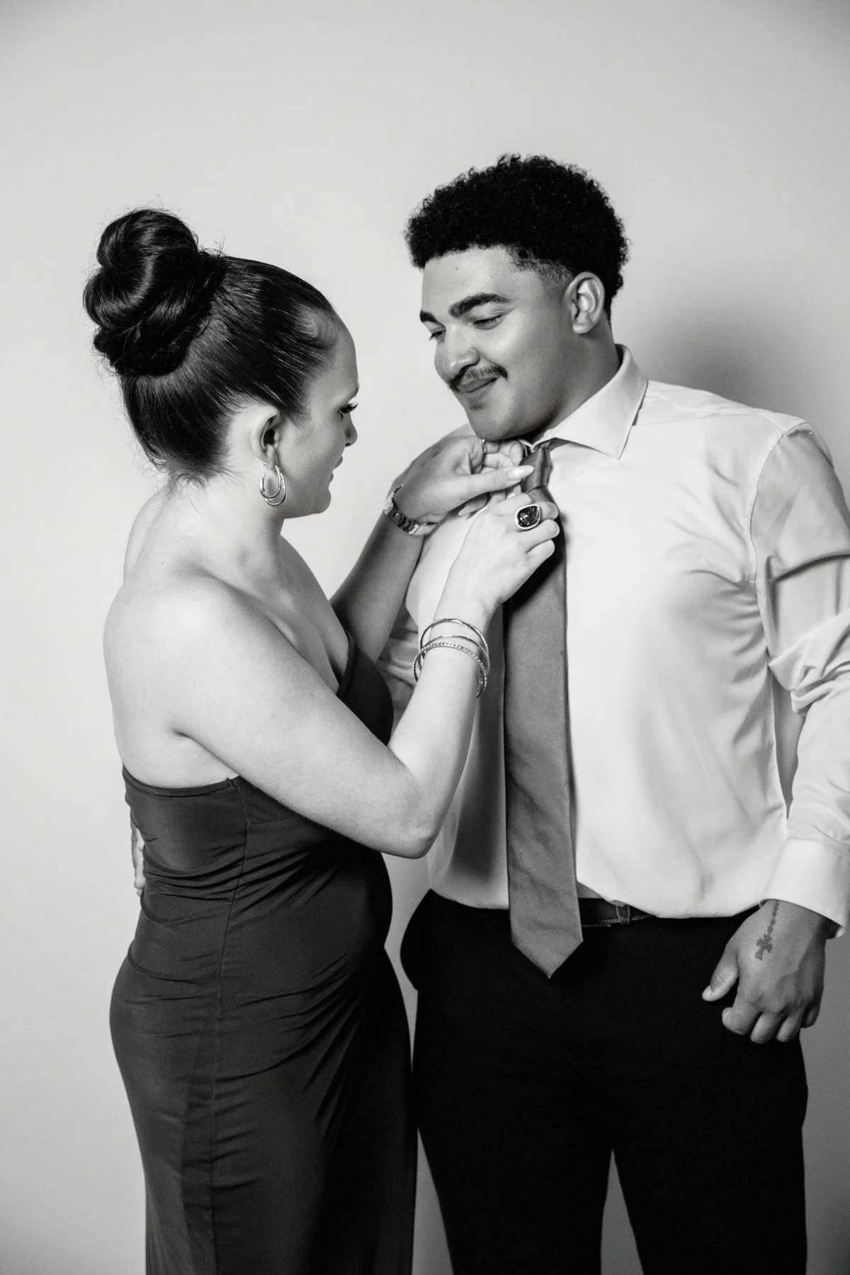 Black and white photo of a woman adjusting a man's tie. The woman has her hair in an updo and is wearing earrings and a sleeveless dress. The man has short curly hair and is wearing a dress shirt and tie, with a ring on his finger and tattoos on his 