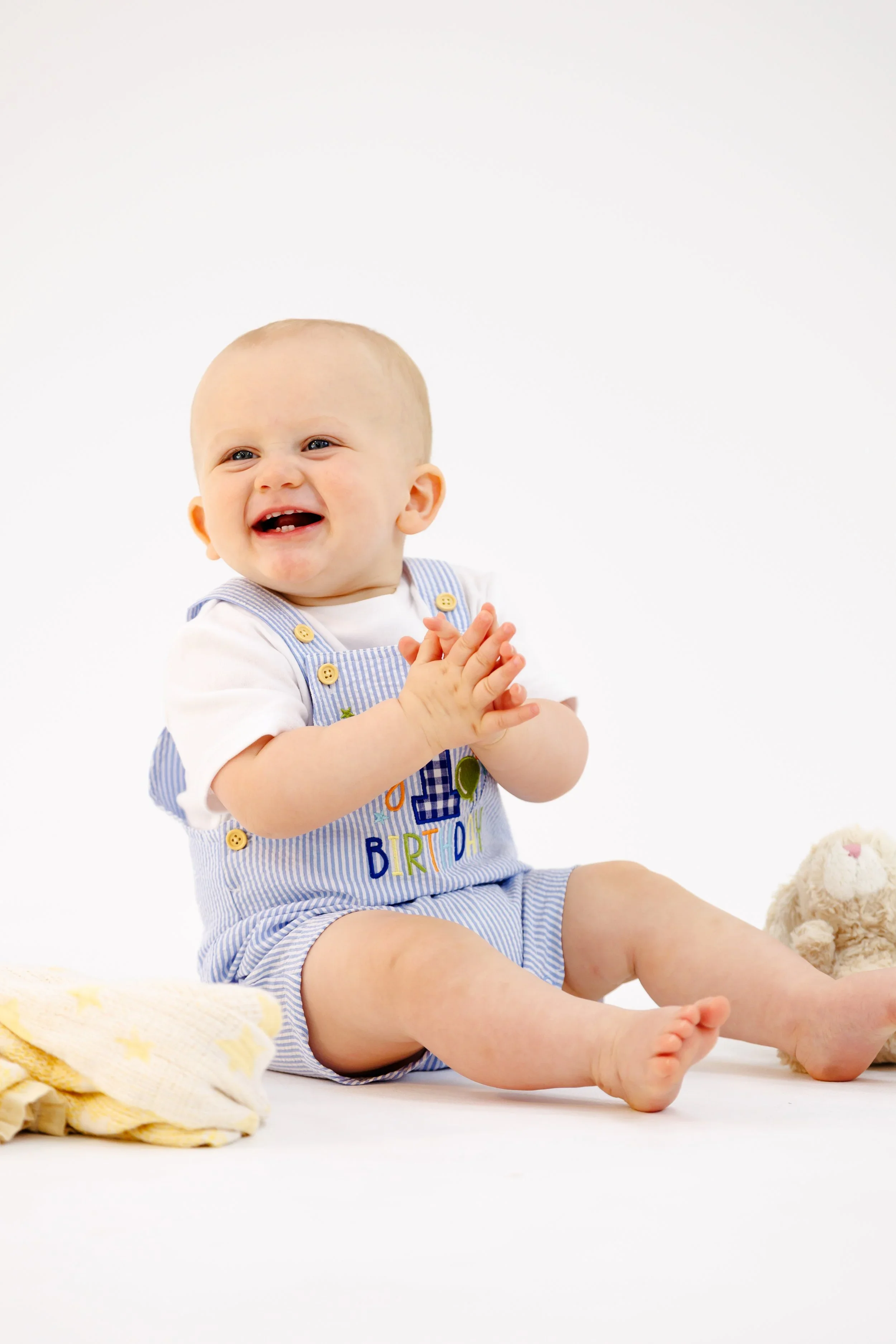 A smiling baby wearing a striped blue and white outfit with a 'Happy Birthday' message, sitting on a white background with a plush teddy bear beside him and a yellow blanket in the foreground.