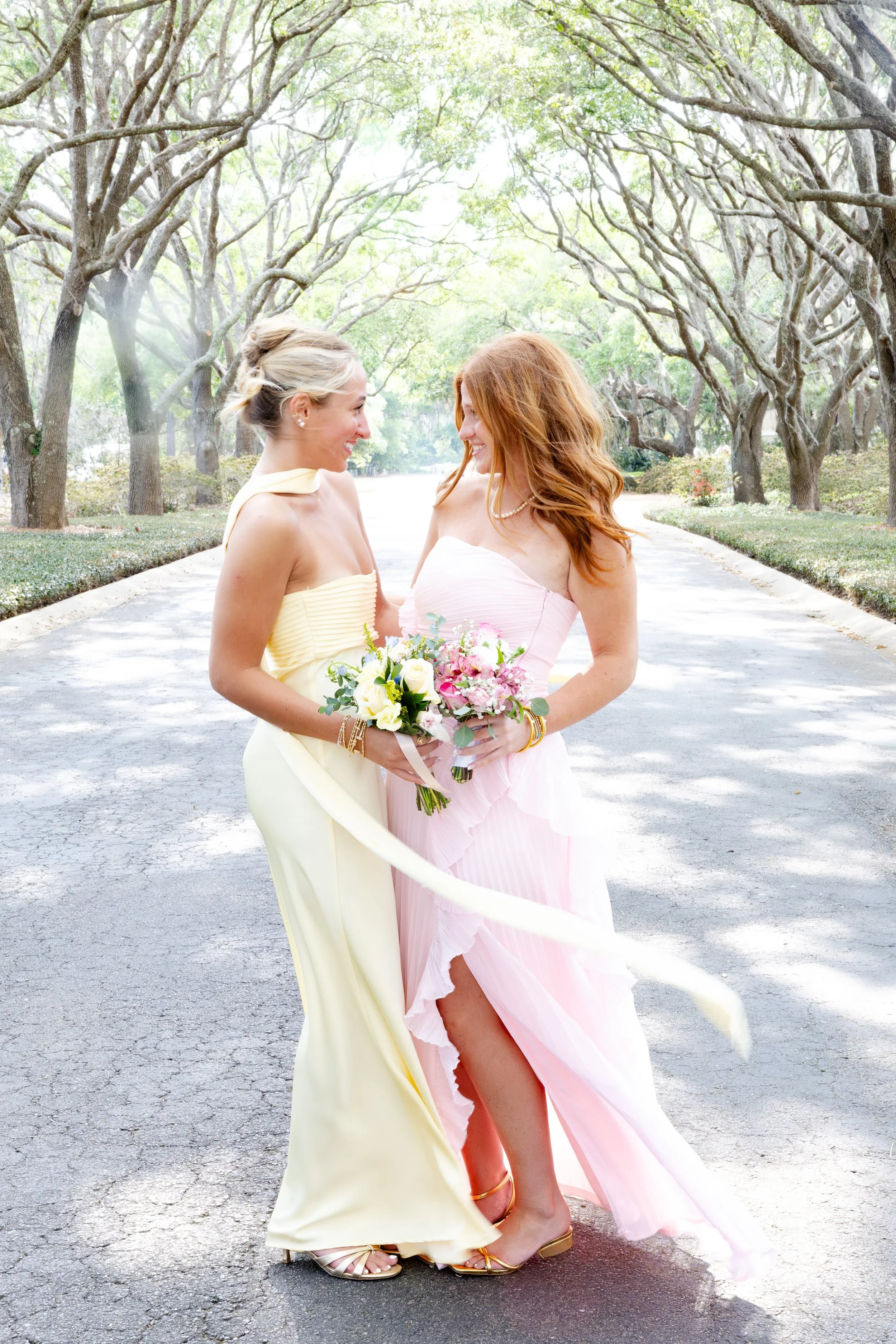 Two women in colorful dresses stand close together on a tree-lined street, smiling and holding bouquets of flowers during the daytime.