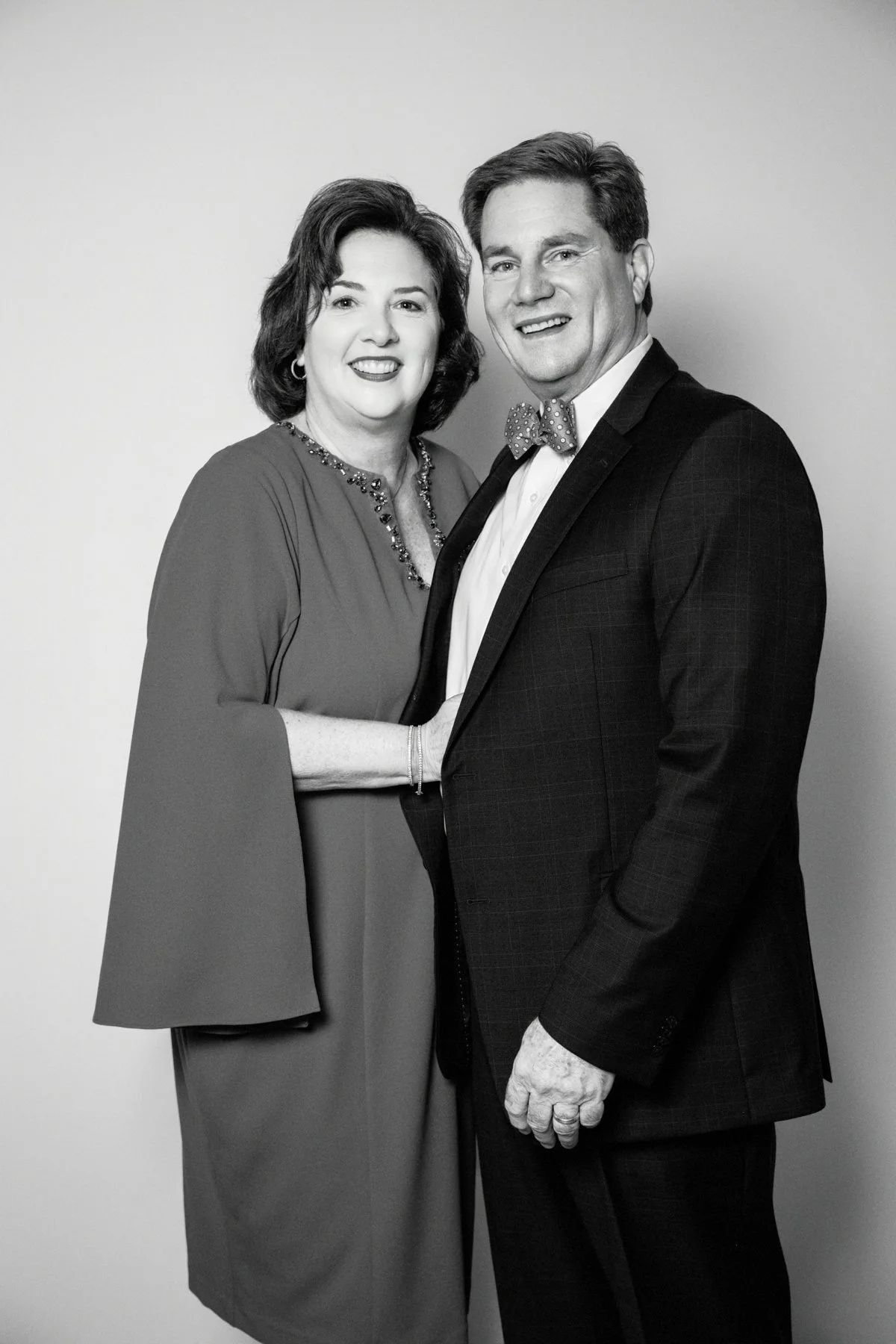 A black-and-white photograph of a smiling man and woman standing close together, with the woman’s hand resting on the man’s chest. The woman has shoulder-length dark hair and is wearing a dress with embellished neckline, while the man has short hair 
