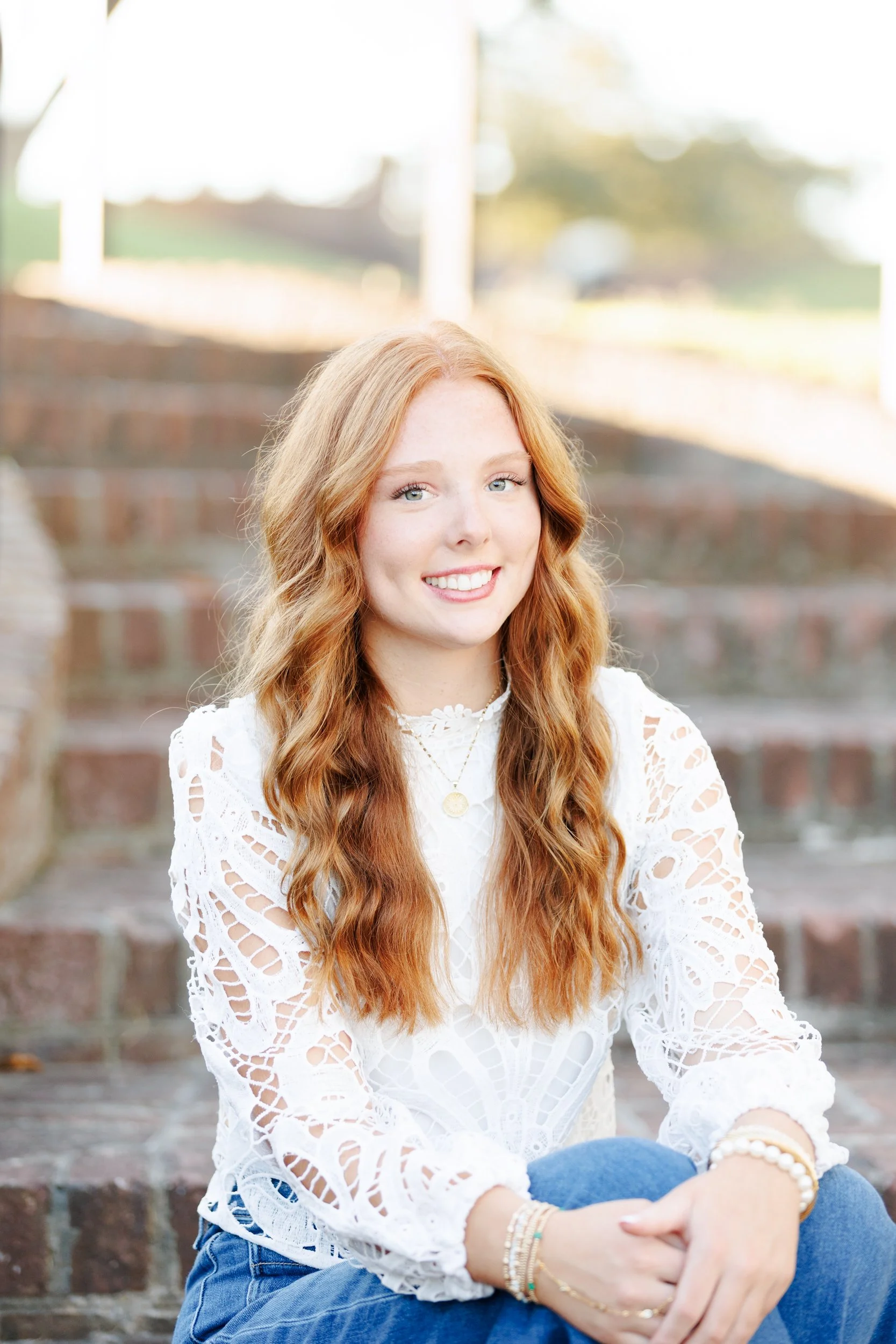 Portrait of a young woman with long red hair sitting on brick stairs outdoors, smiling at the camera, wearing a white lace top and blue jeans.