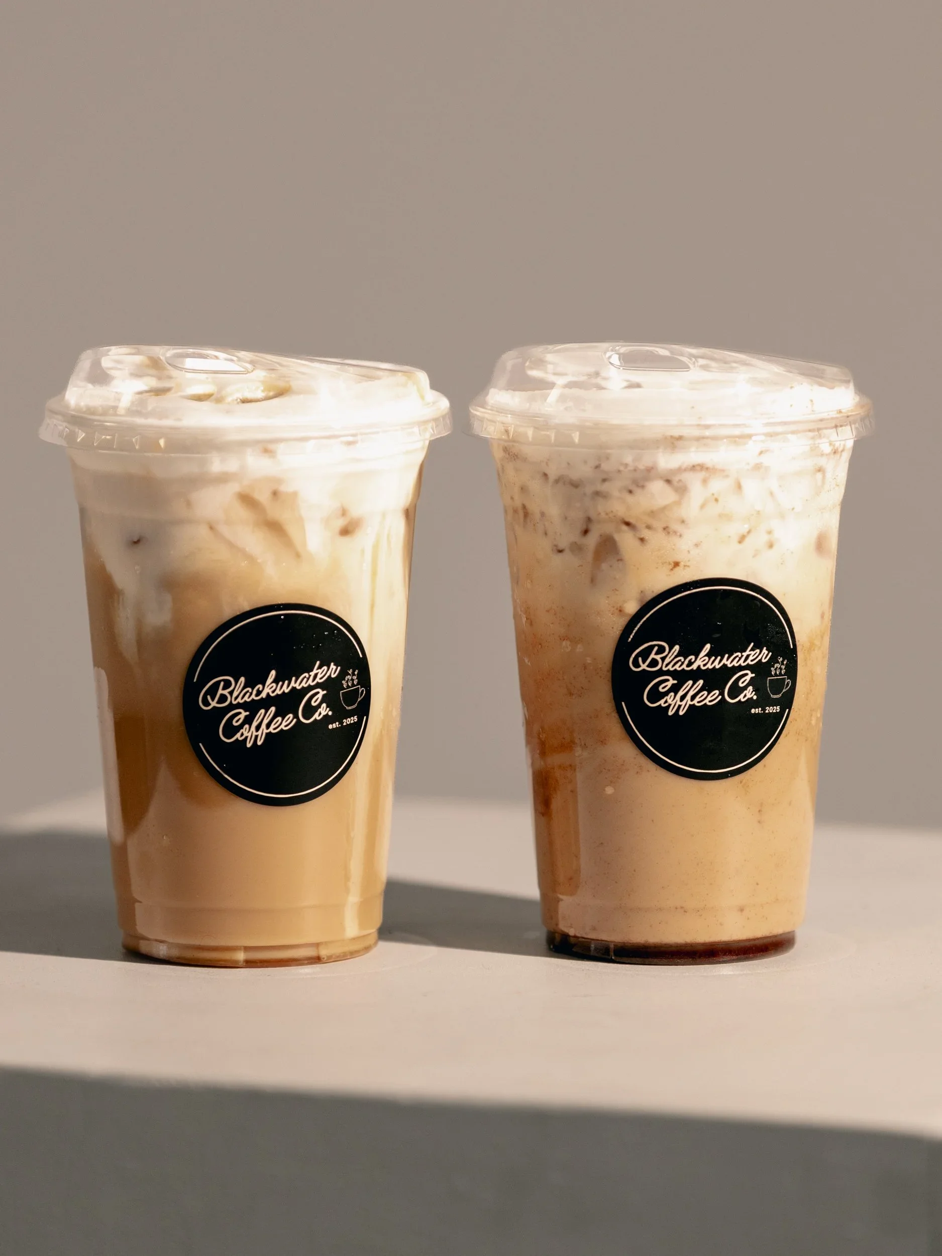 Two iced coffee drinks in clear cups with plastic lids, placed on a white surface against a plain background. The cups have a black circle logo that says "Blackwater Coffee Co." with a small coffee cup icon and "est. 2025".