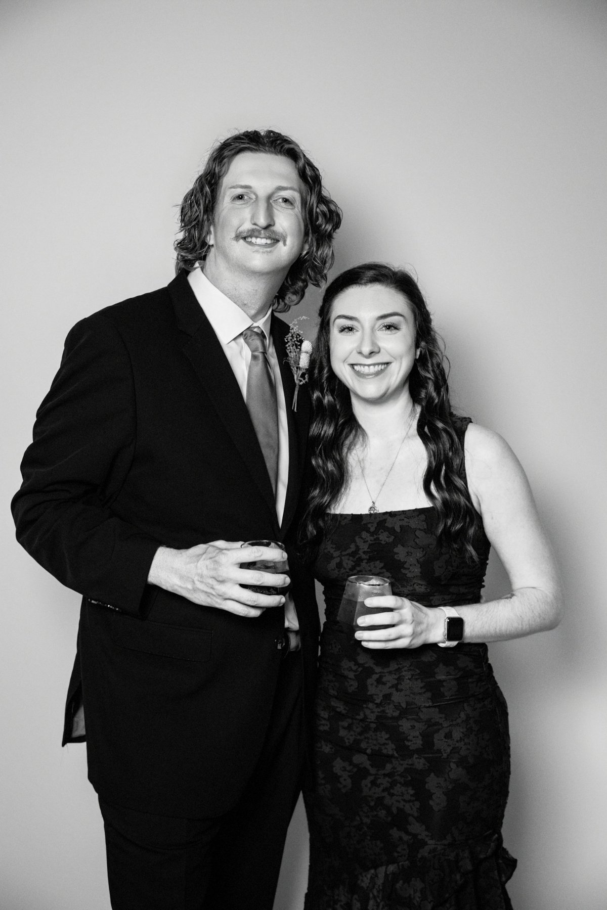 Black and white photo of a man in a suit with long hair and a woman in a dress with long curly hair, both smiling, holding drinks, standing against a plain wall.