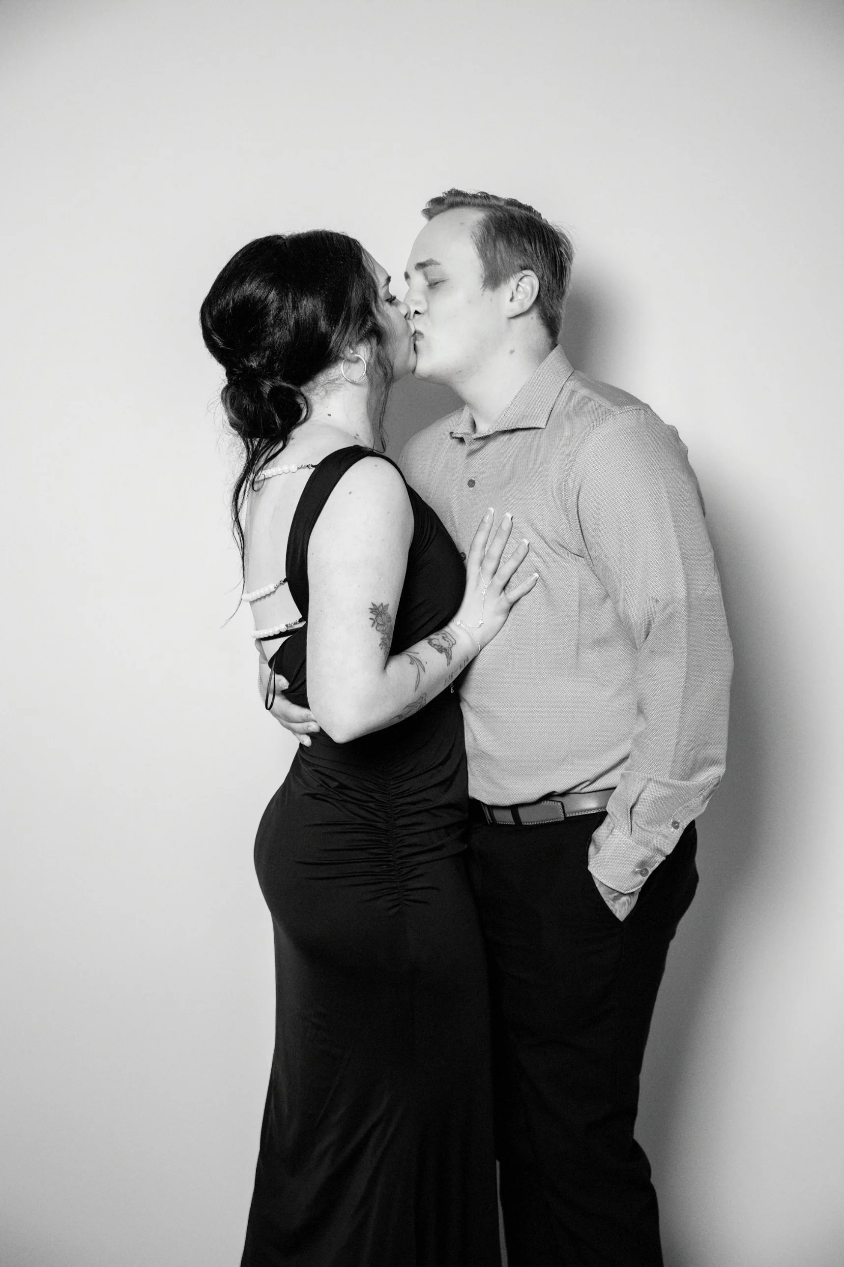 A couple sharing a kiss, with the woman's hand on the man's chest. The woman is wearing a black dress with pearl accessories and has tattoos on her arm. The man is dressed in a light-colored shirt and dark pants, standing against a plain background.