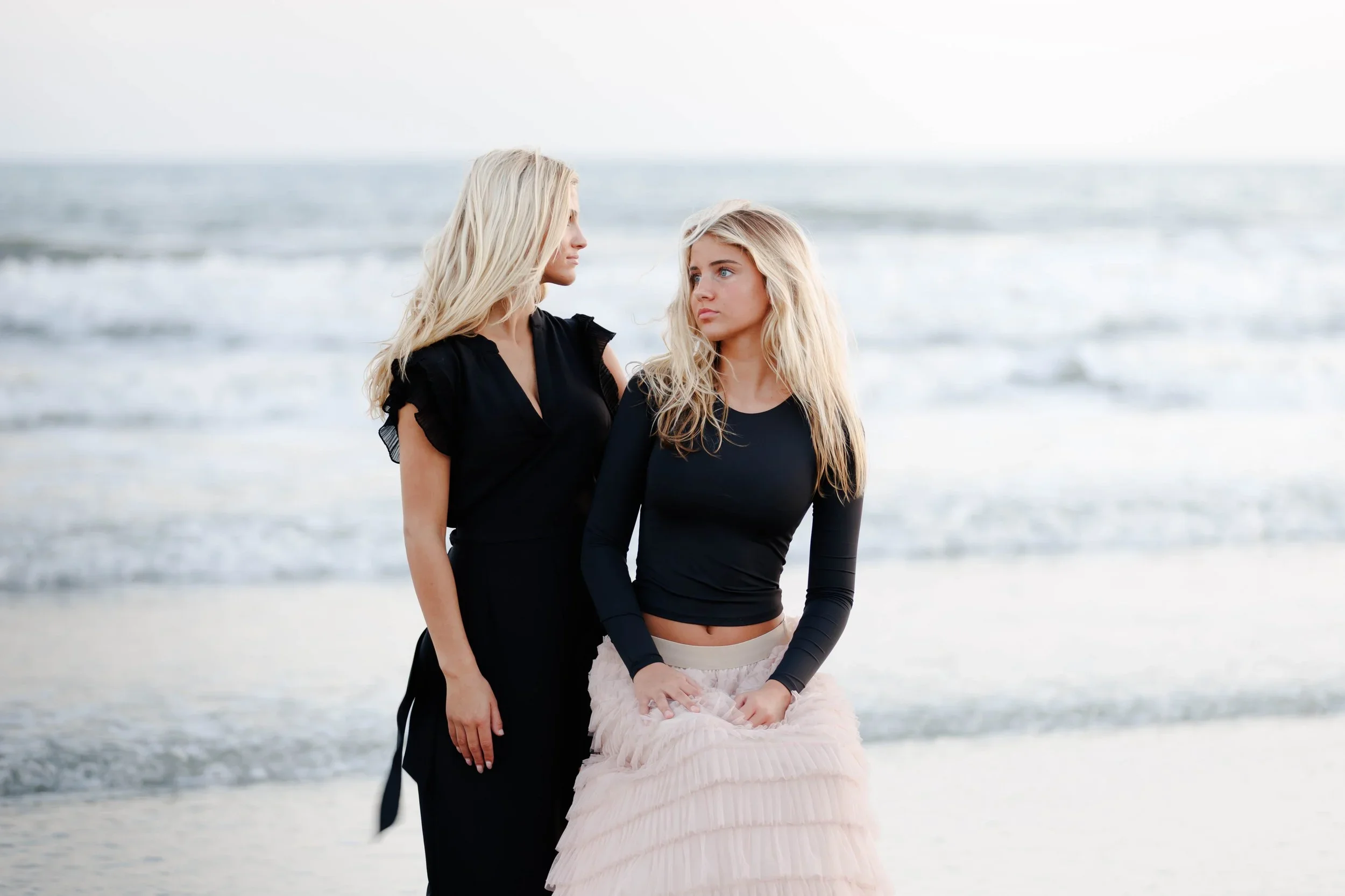 Two women with blonde hair stand on a beach near the ocean, wearing black tops and looking at each other.