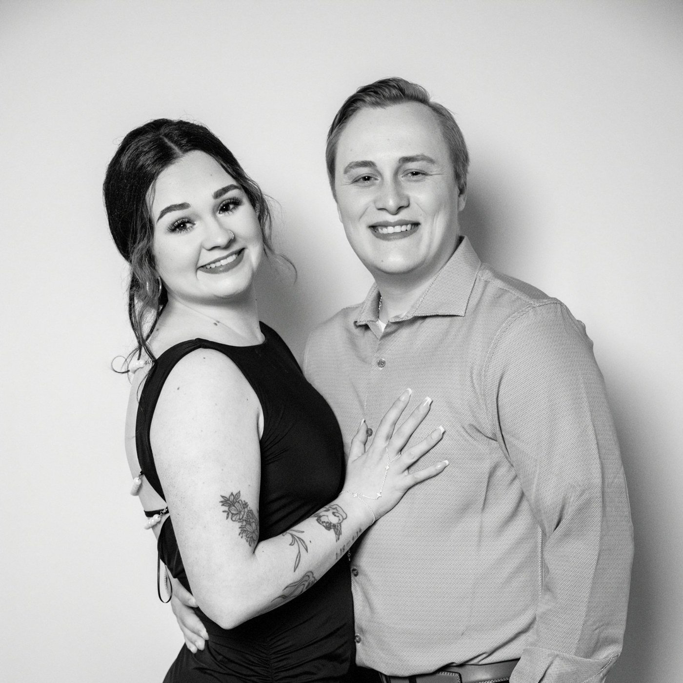 A black and white photo of a smiling couple, a woman with long dark hair and tattoos on her arm, wearing a sleeveless dress, and a man with short light hair, wearing a button-up shirt. They are close together, with the woman holding her hand on the man's stomach.