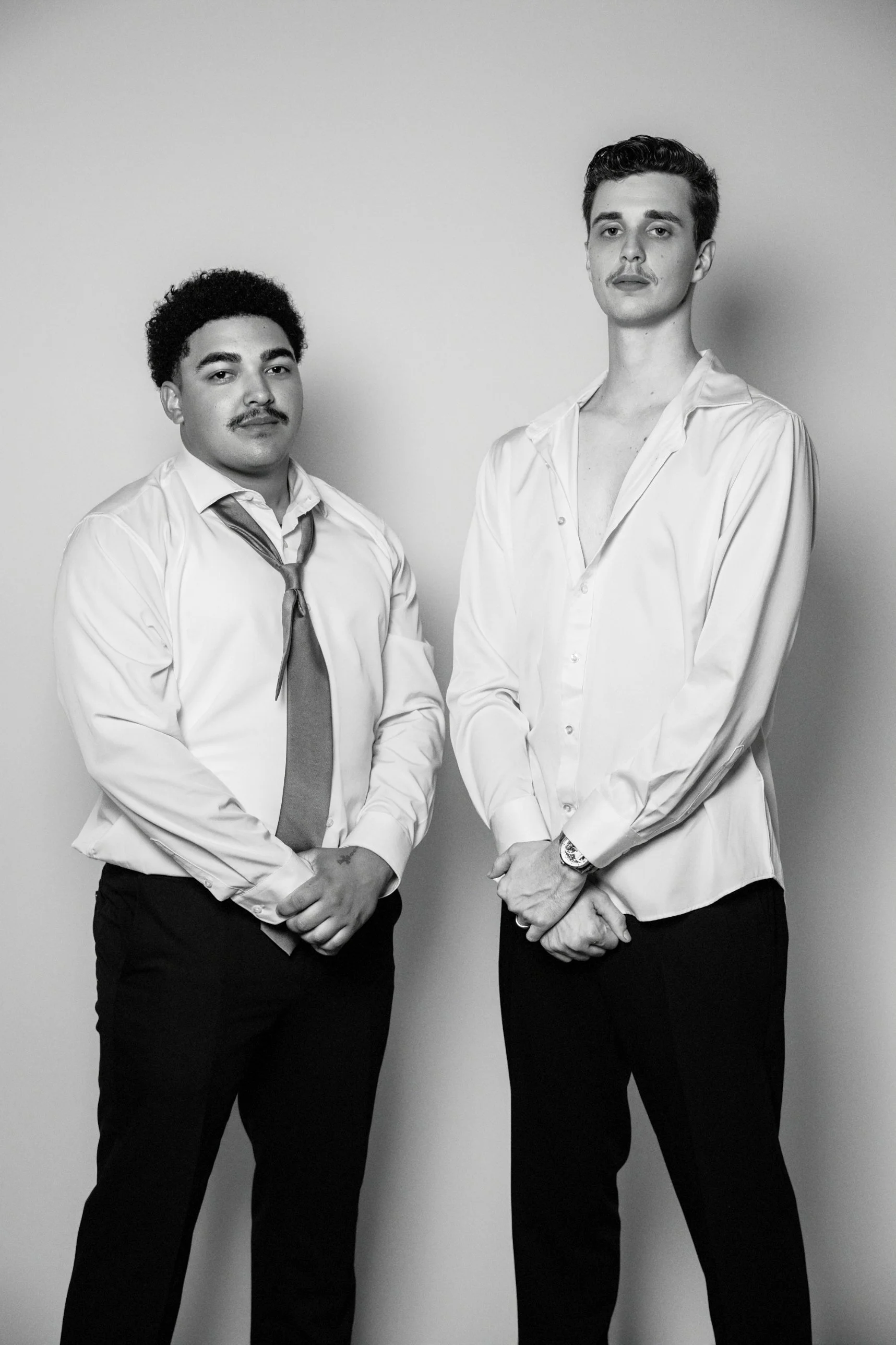 Two young men in white dress shirts and black pants standing against a plain wall, posing confidently for a black-and-white photograph.