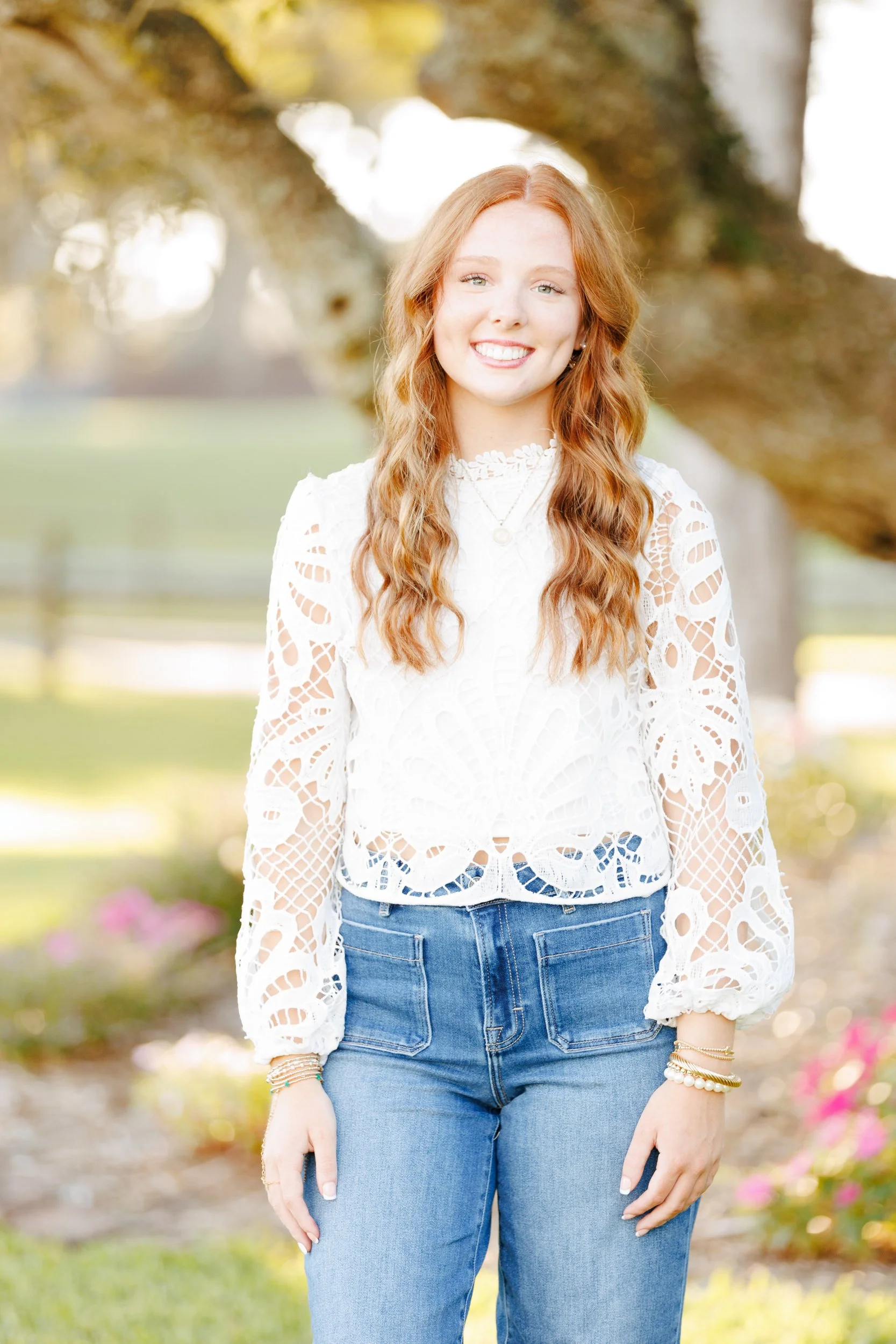 Young woman with long red hair smiling outdoors. She wears a white lace long-sleeve top, blue jeans, and multiple bracelets. The background shows a tree and a park with pink flowers on the ground.
