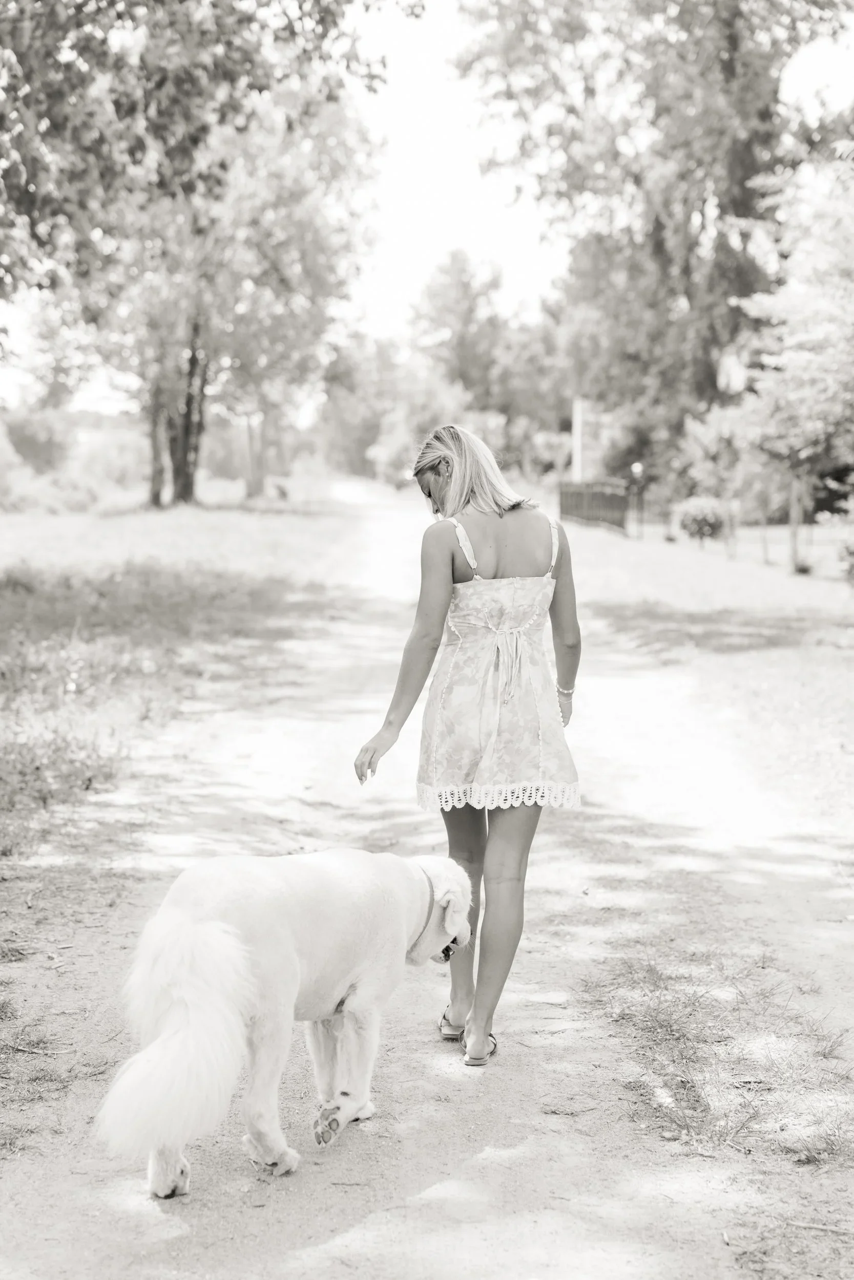 A woman with blonde hair wearing a light-colored dress walking with a large white dog along a dirt path lined with trees in a park.