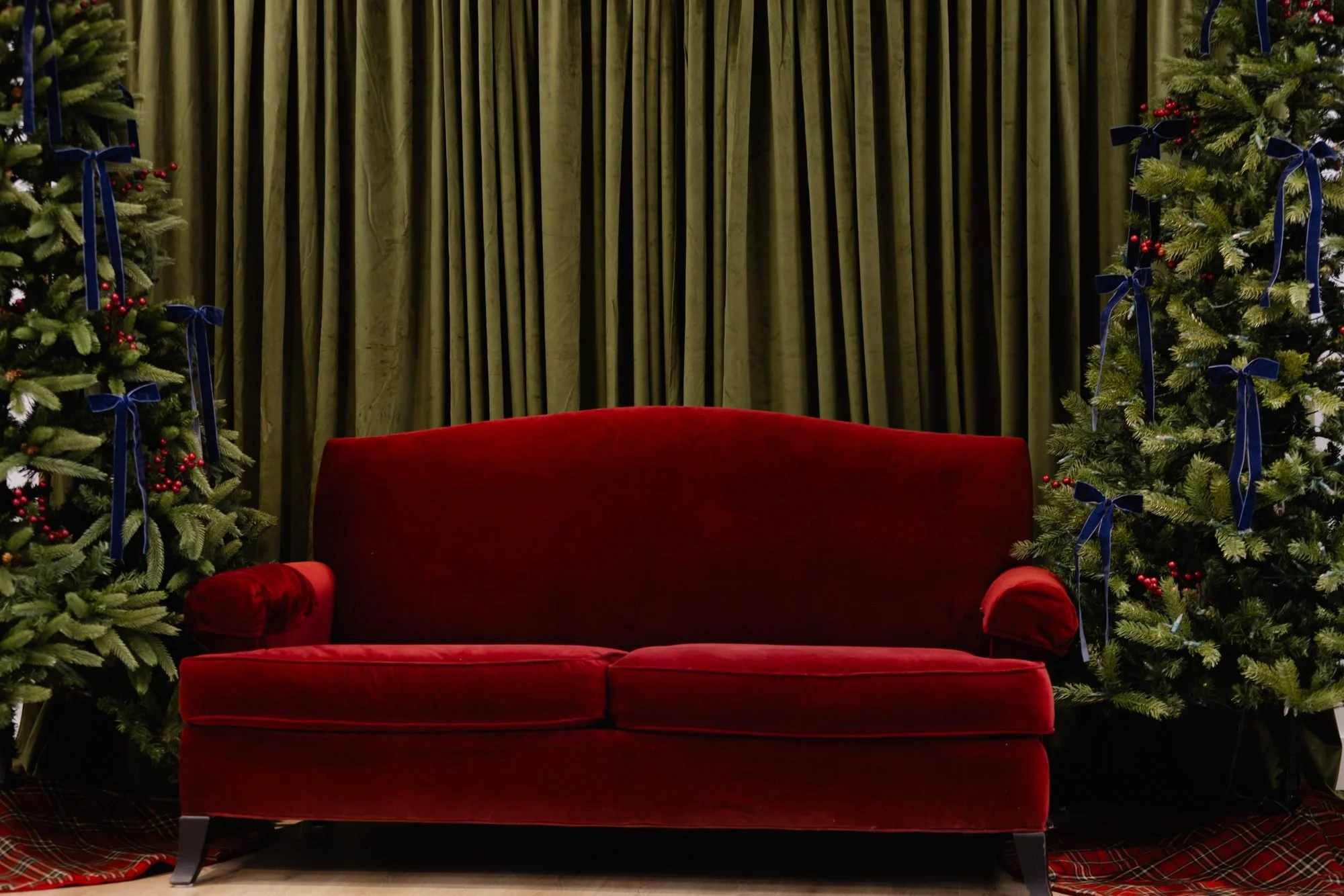 Red velvet sofa in front of green curtain with decorated Christmas trees on each side, featuring blue ribbons and red berries.