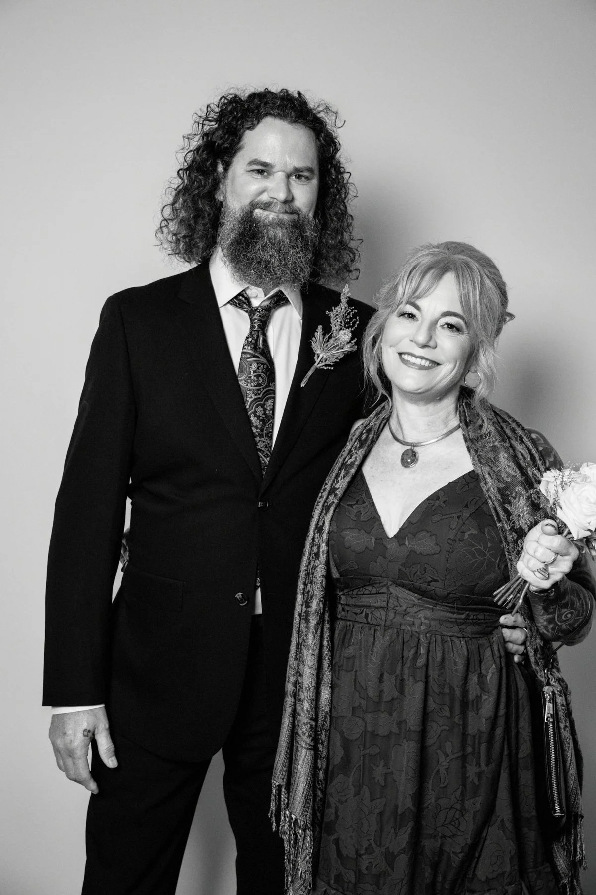A black and white photo of a man with curly hair and a beard wearing a suit with a boutonniere and a woman with short hair holding a flower, both smiling and standing close together against a plain background.
