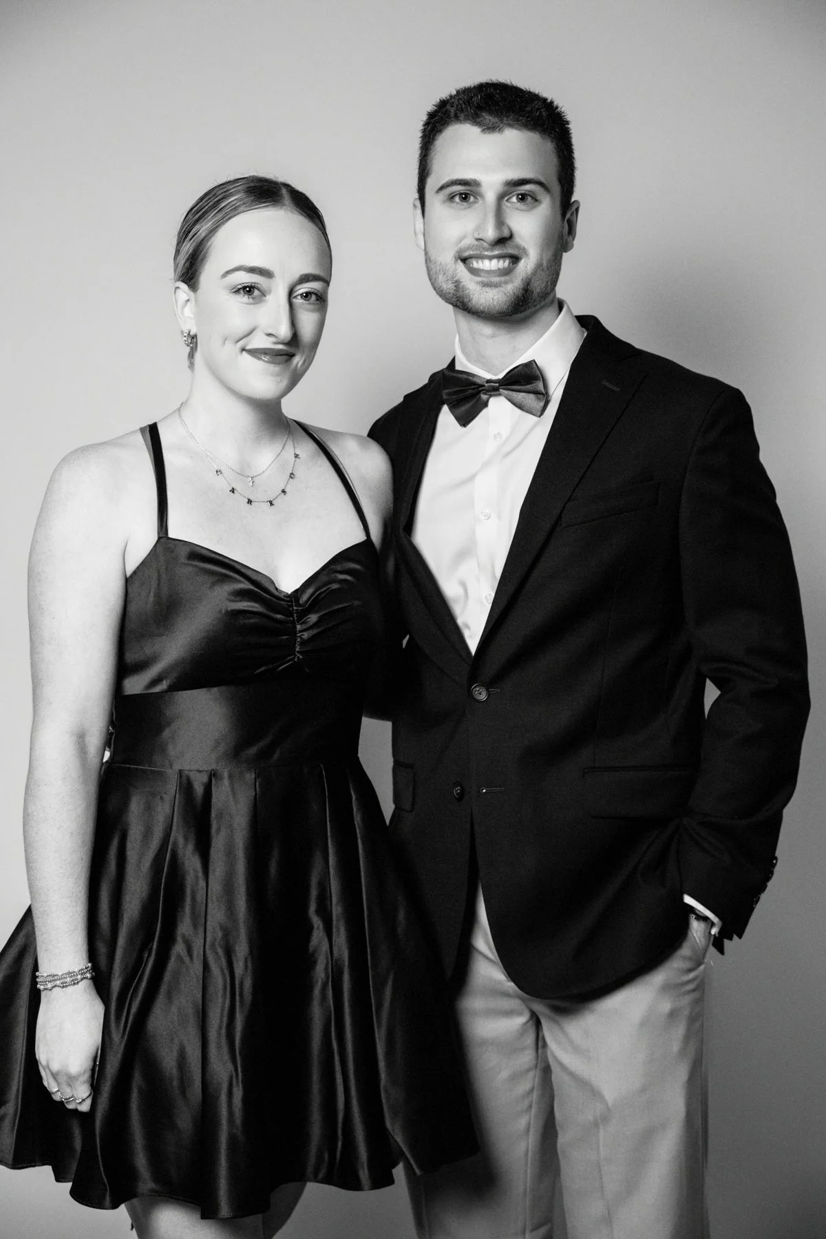 A black-and-white photo of a young woman and man dressed in formal attire, smiling at the camera. The woman is wearing a satin dress with spaghetti straps and layered necklaces. The man is in a tuxedo with a bow tie and a white shirt, with his left h
