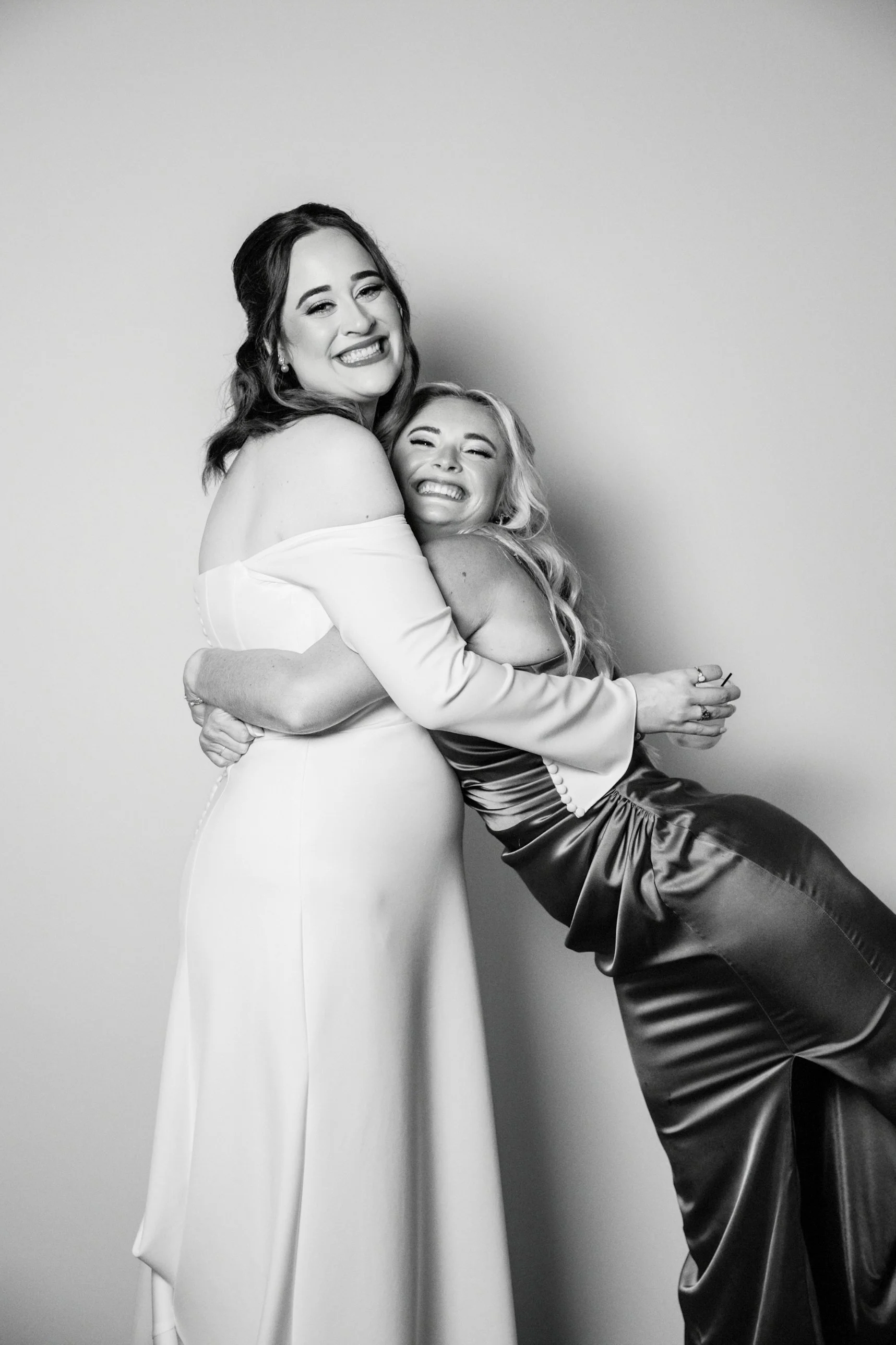 Two women hugging, smiling happily, one in a white dress, the other in a satin dress, black and white photograph.