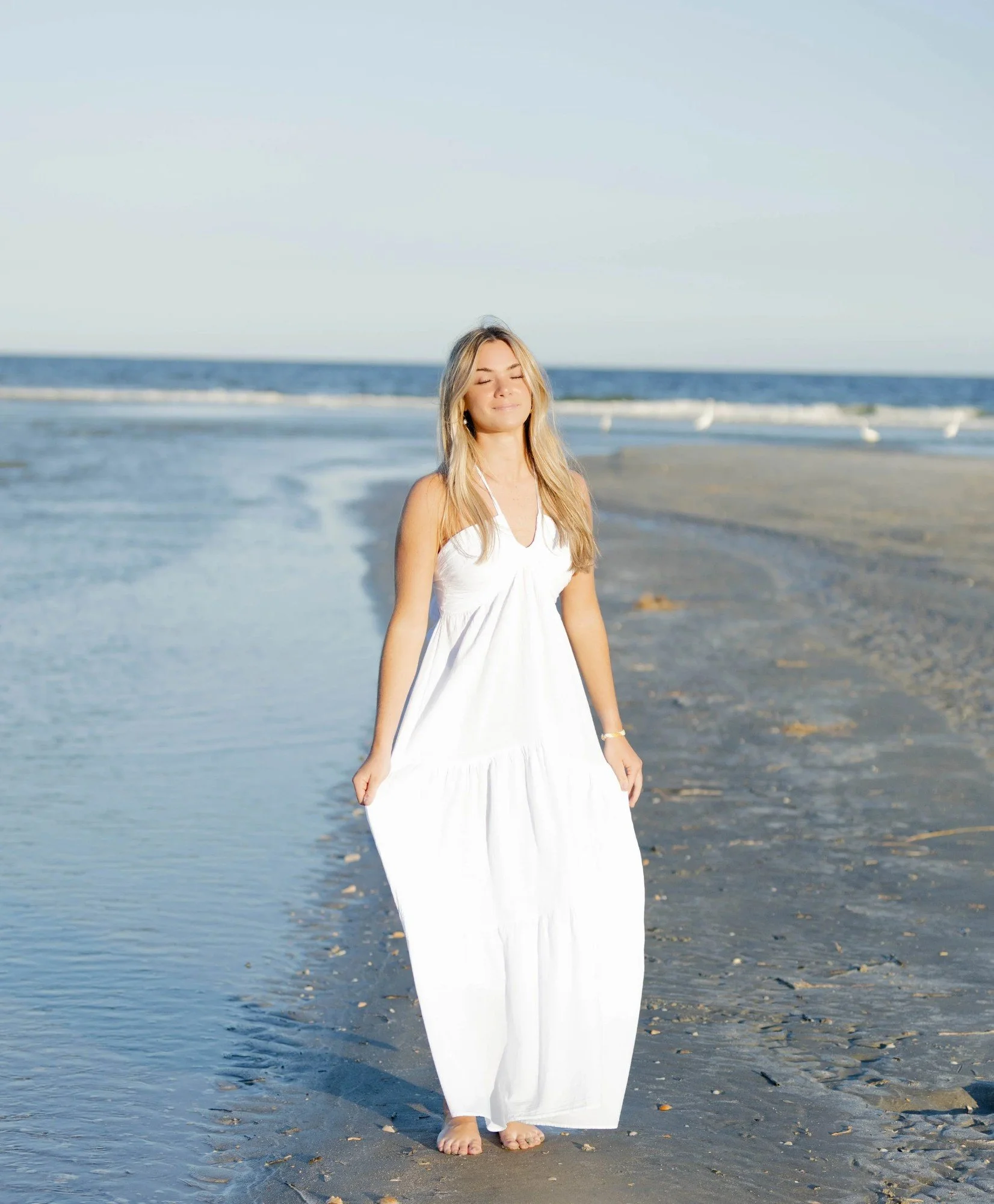 A woman in a white dress with long blonde hair standing barefoot on a sandy beach near the ocean, with her eyes closed and a peaceful expression.