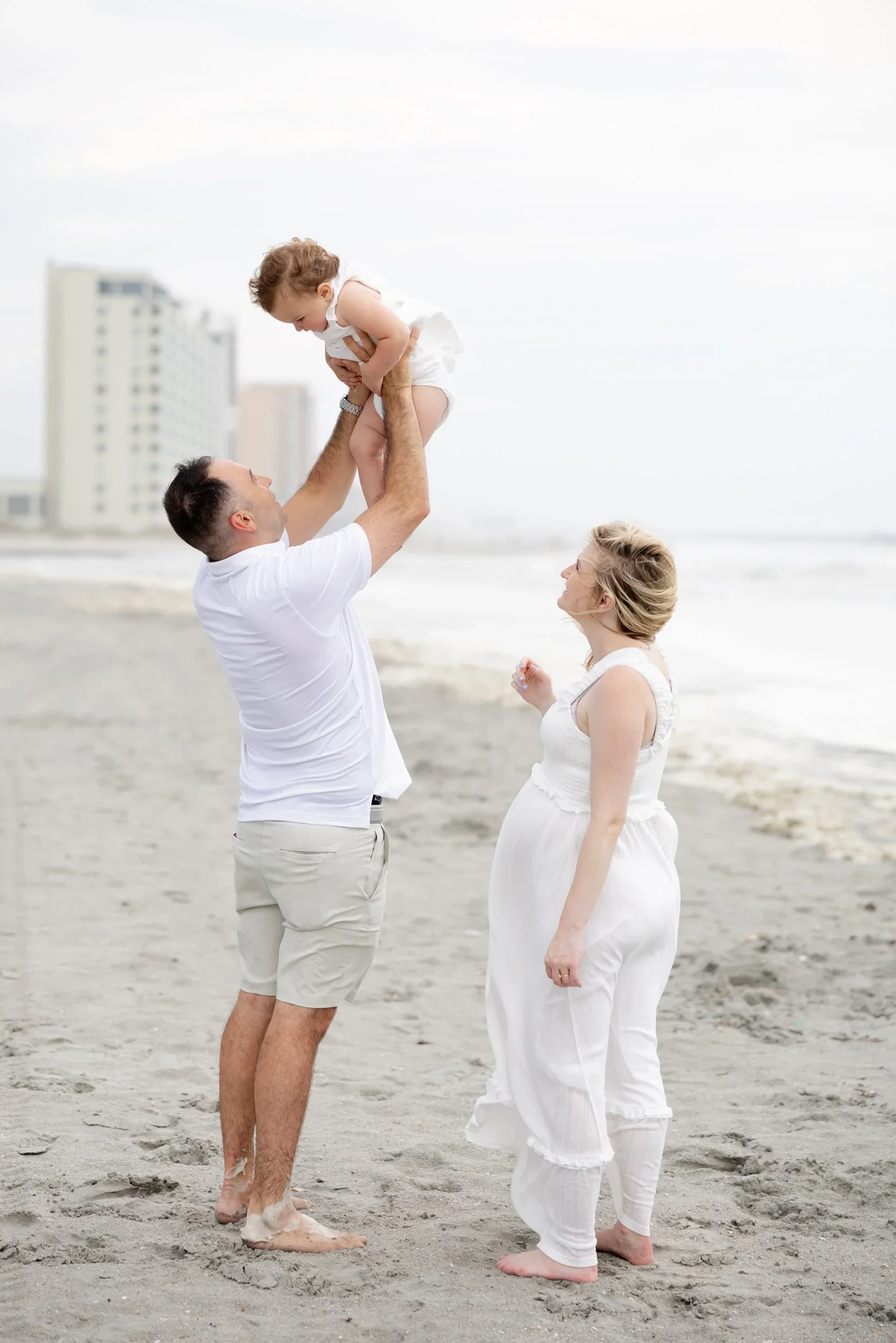 A man holds a young girl up in the air on a beach, with a woman watching and smiling. The background features tall buildings and a cloudy sky.