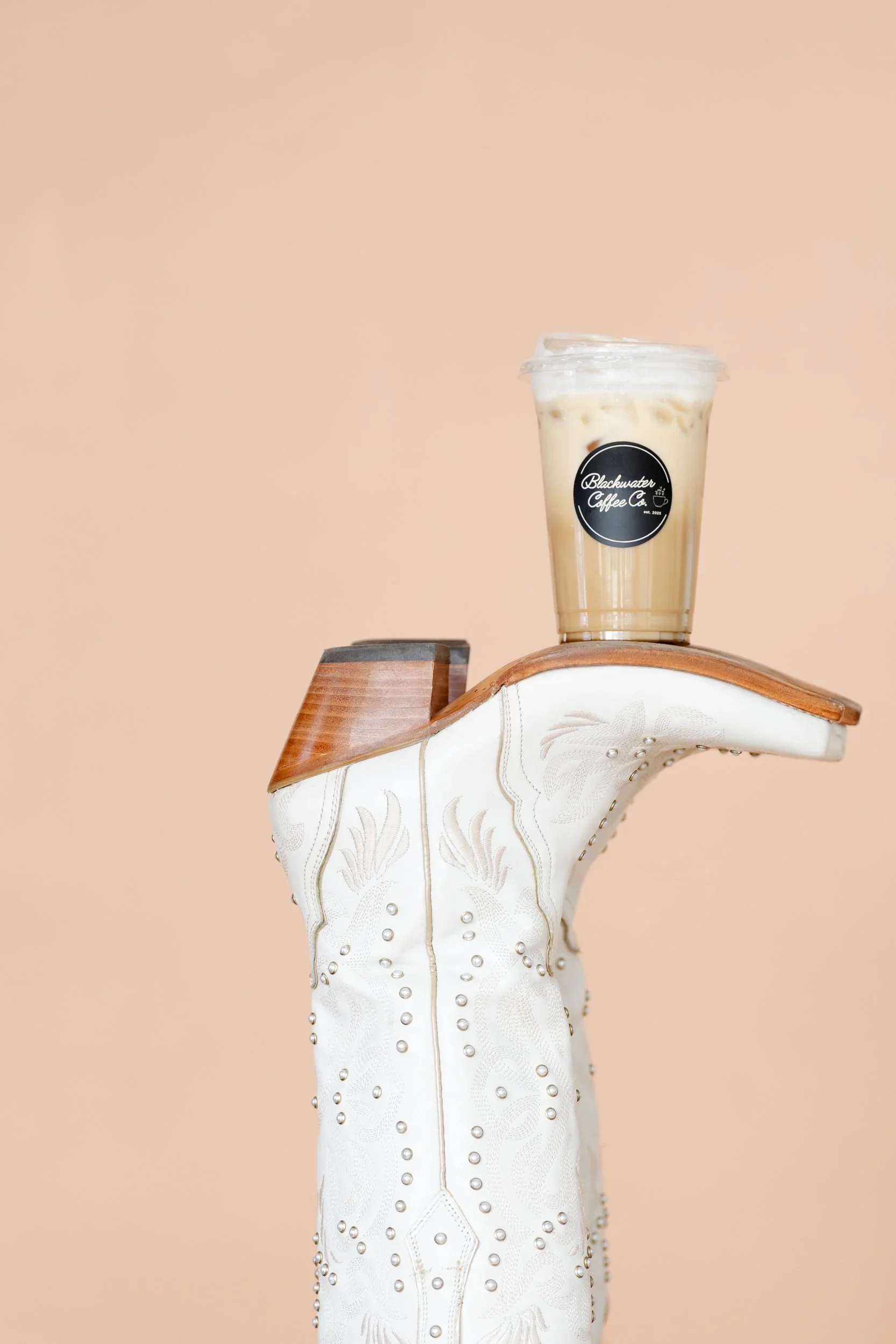 Glass of iced coffee with milk and a black logo sticker from Blackwater Coffee Co., placed on a decorative white and wooden surface against a beige background.