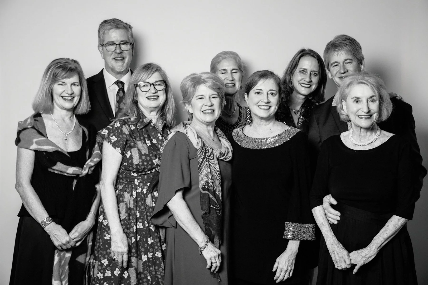 Black and white photo of eleven women and one man standing together against a plain wall. They are all smiling and dressed in formal attire, with some wearing jewelry and patterned clothing.