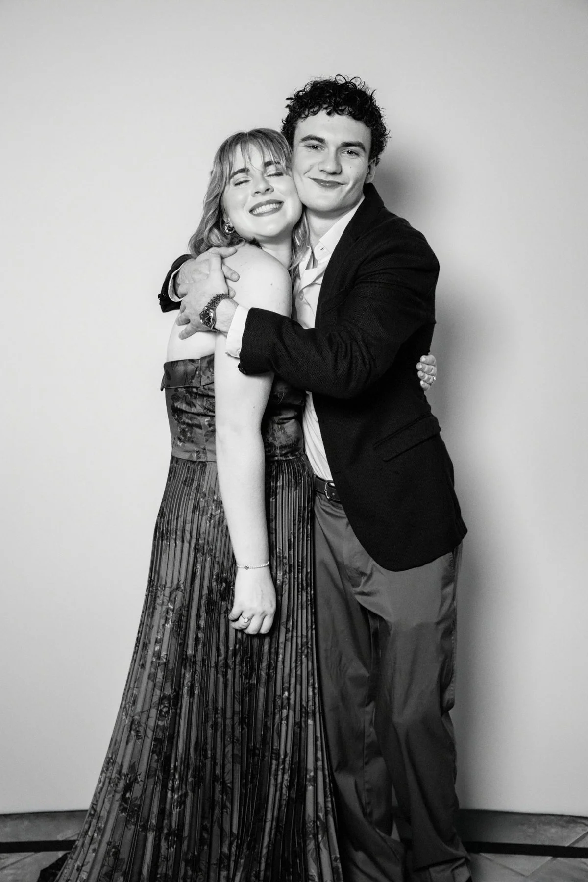 A black and white photo of a woman and a man hugging, both smiling. The woman has shoulder-length hair, is wearing a sleeveless top and a pleated skirt with a floral pattern, and has makeup on. The man has curly hair, is wearing a suit jacket, dress 