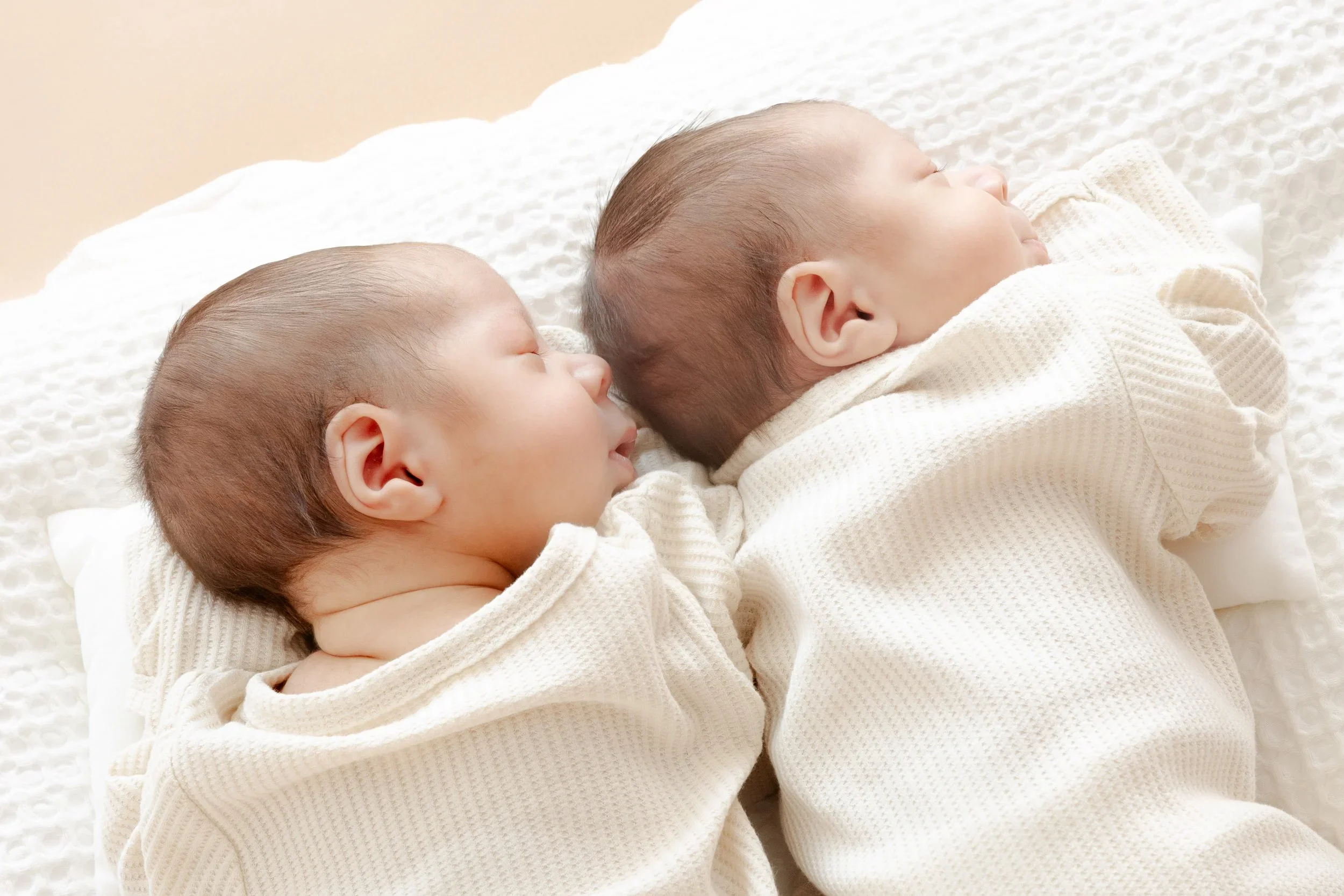 Three sleeping babies lying side by side on a white textured surface, dressed in cream-colored sweaters.