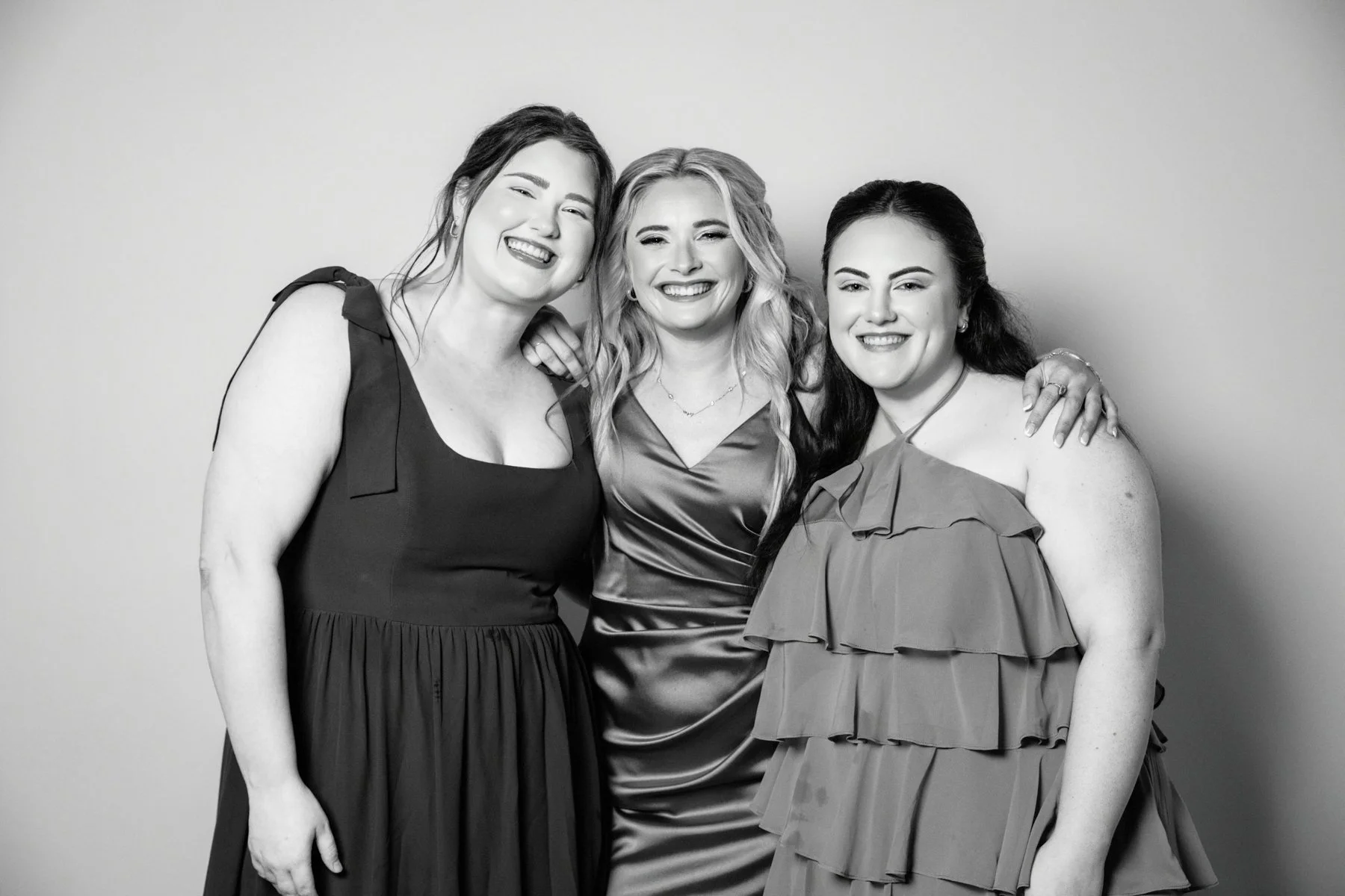 Three women standing together, smiling in front of a plain background. The woman on the left is wearing a sleeveless black dress with a shoulder bow, the woman in the middle is in a shiny satin dress with wavy blonde hair, and the woman on the right 