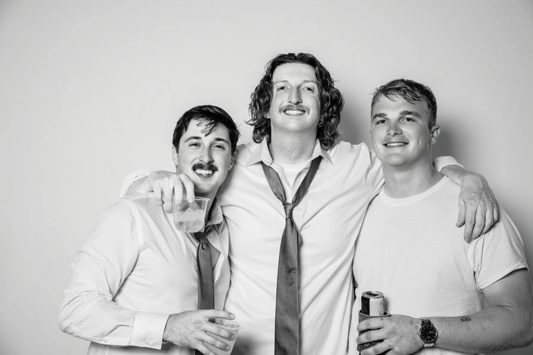 Three men smiling and posing together, with one holding a drink and another holding a can, in a casual setting, in black and white.