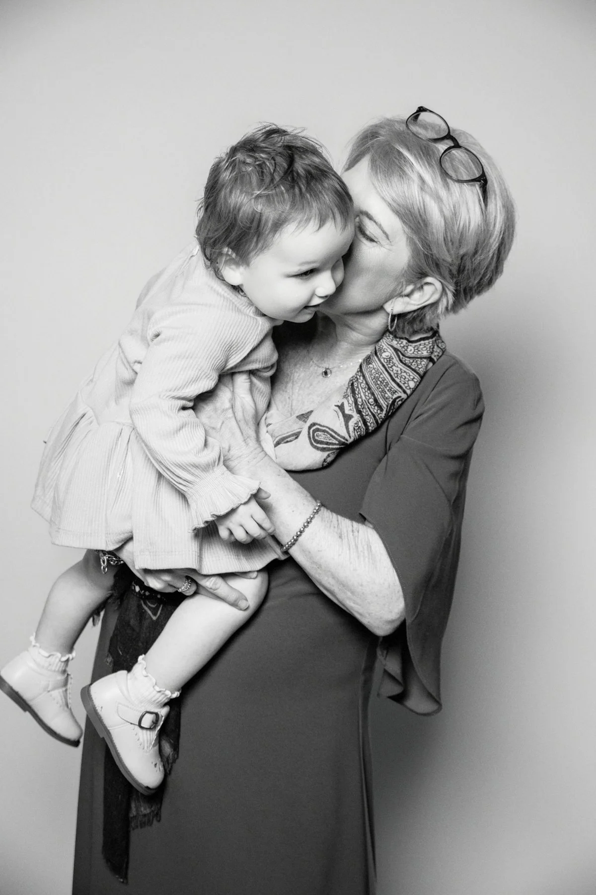 An elderly woman with short hair, earrings, and glasses on her head, holding a young boy close and kissing him on the cheek. The boy has curly hair and is wearing a long-sleeved shirt, shorts, and sneakers. The background is plain.