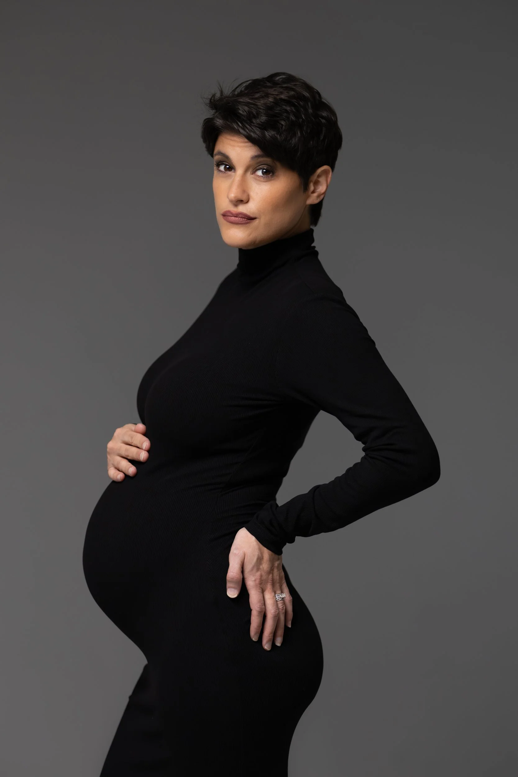 A pregnant woman with short black hair wearing a black turtleneck sweater, standing sideways with her hand on her belly and looking at the camera, against a gray background.
