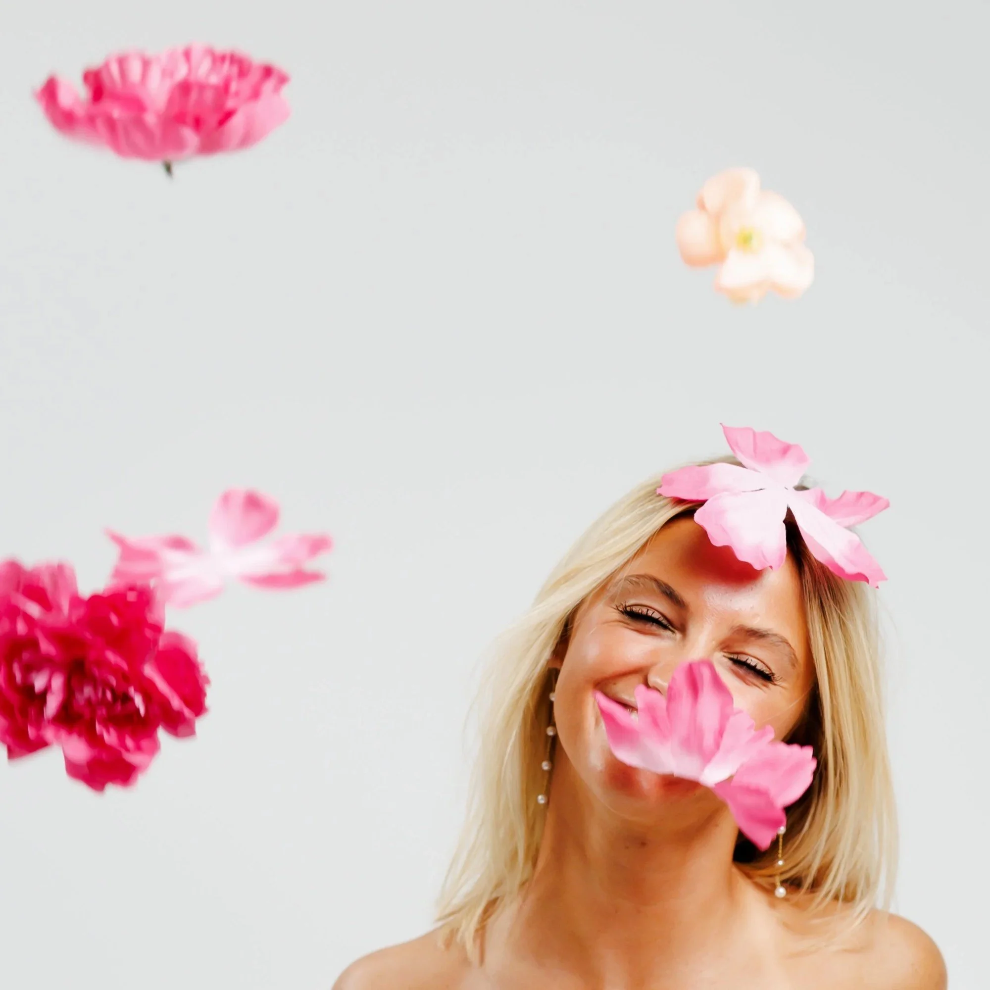 Part of a smiling blonde woman with earrings, decorated with pink flower petals, against a plain white background.